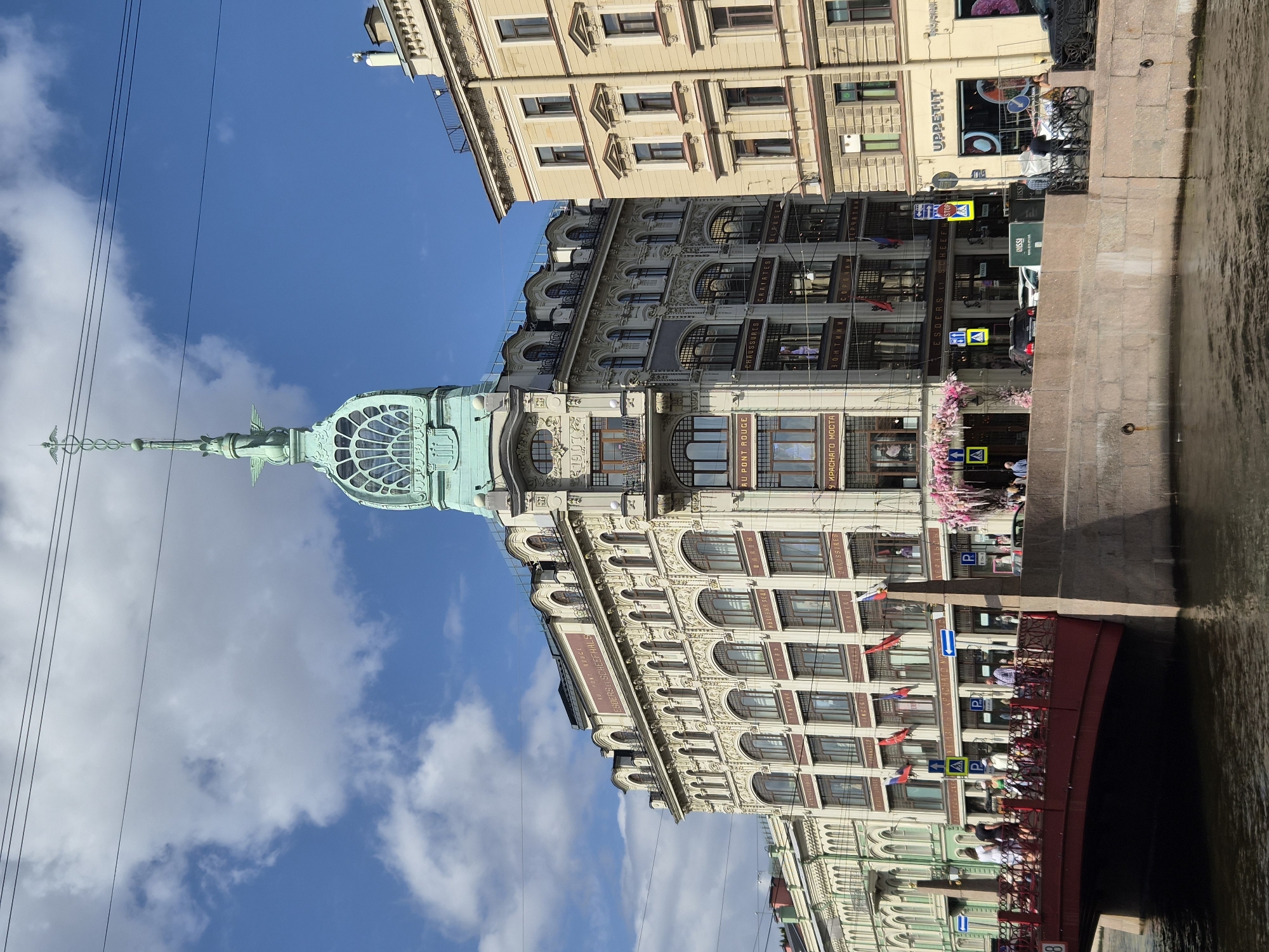An ornate, historic building with a distinctive green domed spire stands prominently on a canal, overlooking a red bridge. The building, a multi-story structure with intricate architectural details and numerous arched windows, displays text in both Russian and French, suggesting it was once a department store. Flags hang from the facade, and pink floral decorations adorn the entrance. A red pedestrian bridge with ornate railings spans the canal, with several people walking across it. The water of the canal reflects the sky and the surrounding architecture. The sky is blue with scattered white clouds. Adjacent buildings, one a lighter yellow color with classical detailing, are also visible. Various signs, including traffic and parking signs, are present. Vehicles, including a black car and a white car, are parked along the canal. The overall scene depicts a sunny day in a picturesque European city.