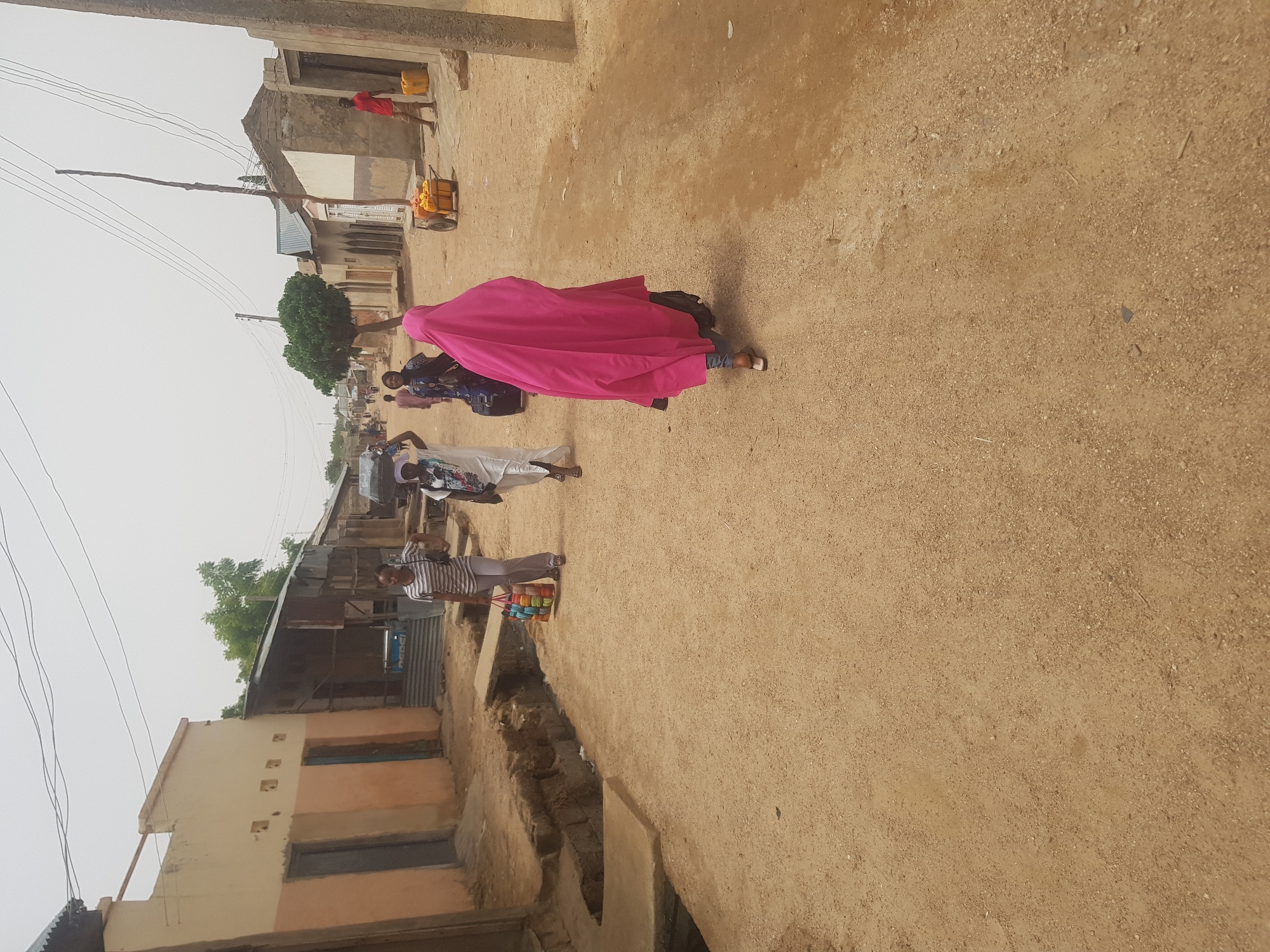 A street scene in Dutsinma B, Nigeria, shows several people walking on a dirt road lined with buildings. A woman in a bright pink hijab is prominently featured in the middle ground. Other people are wearing lighter clothing, including white and striped patterns. One person carries colorful items. Buildings are a mix of finished and unfinished construction, with utility poles and overhead wires. The ground is dusty and dry. A drainage ditch runs along the side of the road. In the background, a man pulls a cart loaded with yellow containers.