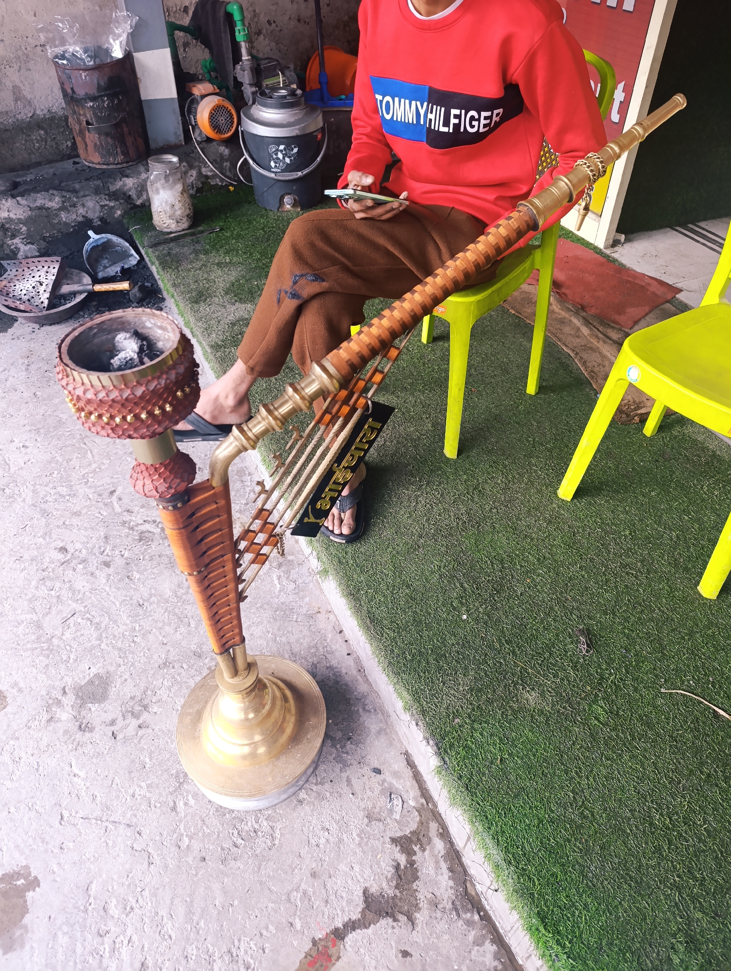 A person wearing a red Tommy Hilfiger sweatshirt and brown pants sits on a bright green plastic chair. They are holding a smartphone in their left hand. In front of them is a large, ornate hookah with a brass base and a decorative wooden stem. The hookah is placed on a concrete floor, next to an area covered with artificial grass. Various objects are visible in the background, including a metal container with a plastic bag inside, a water cooler, and a small orange motor. There is also a sieve and a small shovel with a wooden handle, suggesting preparation for using the hookah. A black license plate with Hindi text is attached to the side of the hookah. The setting appears to be an outdoor space, possibly a shop or a cafe, with the concrete floor and artificial grass indicating an urban or commercial environment. The lighting suggests it is daytime.
