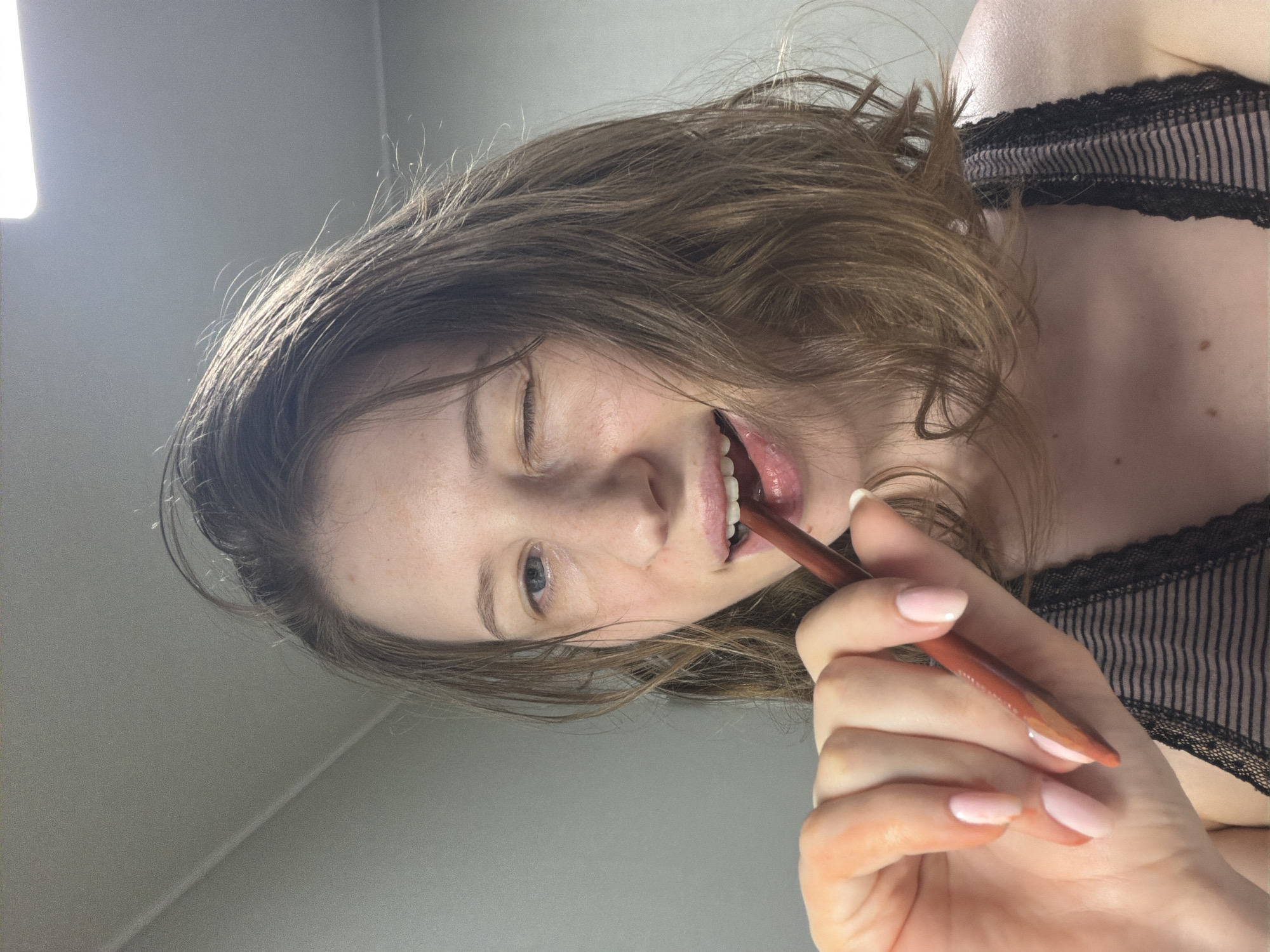 This media file features a close-up, rotated shot of a young Caucasian woman with medium-length brown hair. She is looking directly at the camera with a playful expression, winking one eye and holding a brown makeup pencil to her lips. Her lips are slightly parted, revealing her teeth, and the pencil appears to be a lip liner or a lipstick. She has light skin with some freckles and is wearing a black lace-trimmed top. Her fingernails are painted a pale pink. The background is a plain, light gray wall, with a bright, diffused light source visible on the upper left side of the frame, suggesting an indoor setting, possibly a bathroom or dressing area. The lighting is soft and even, with no indication of the time of day or weather. The overall mood is informal and lighthearted, possibly a moment of self-care or playful experimentation with makeup. The image was captured in Dongducheon-si, South Korea.