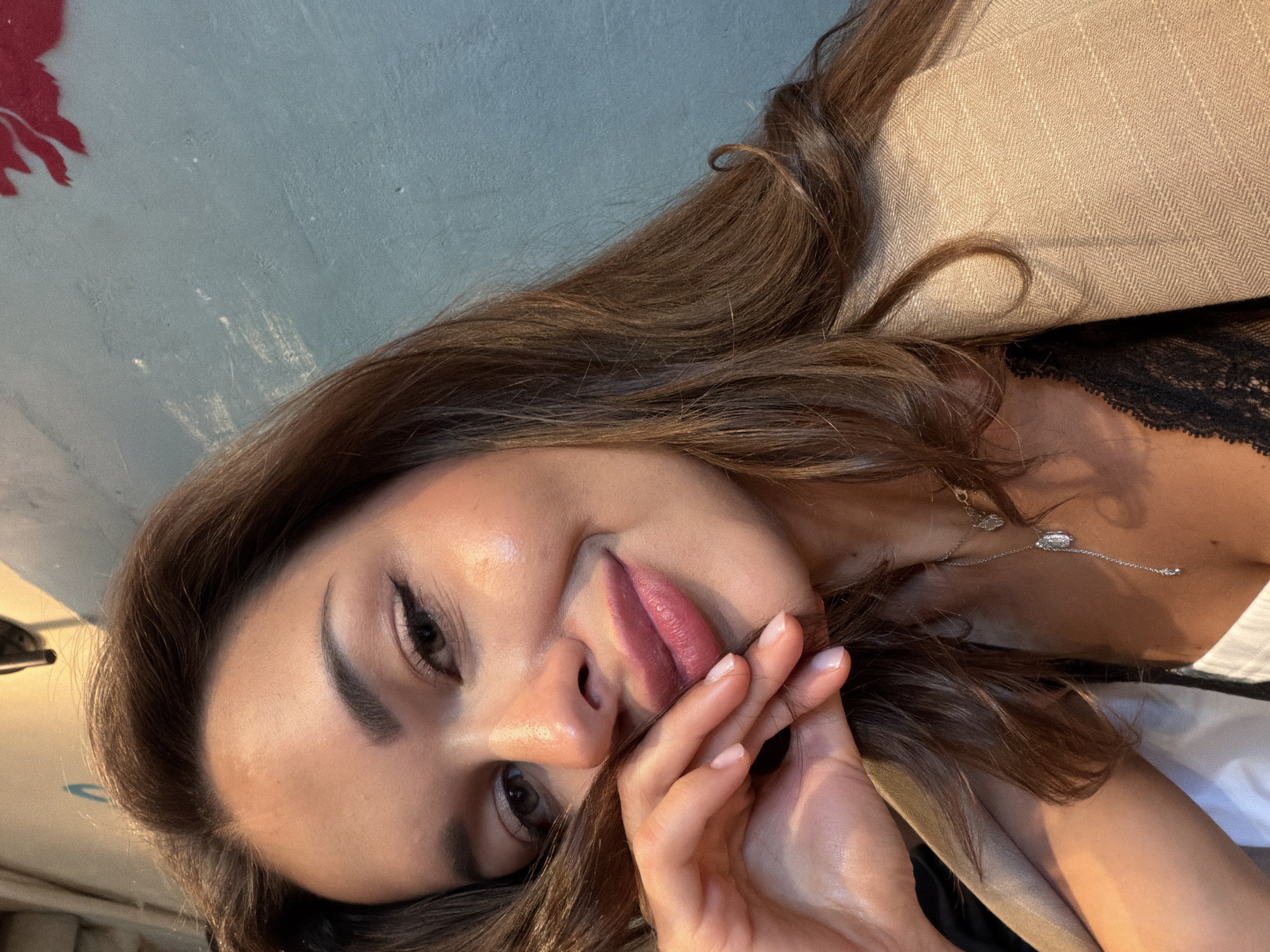 In Saint Petersburg, Russia, a young woman with long, brown hair and fair skin smiles directly at the camera. She is wearing a beige blazer over a black lace top, and a delicate silver necklace with a pendant. Her left hand is placed on her cheek, with her fingers gently touching her lips. The background is a textured, blue-grey wall with some faint white markings.