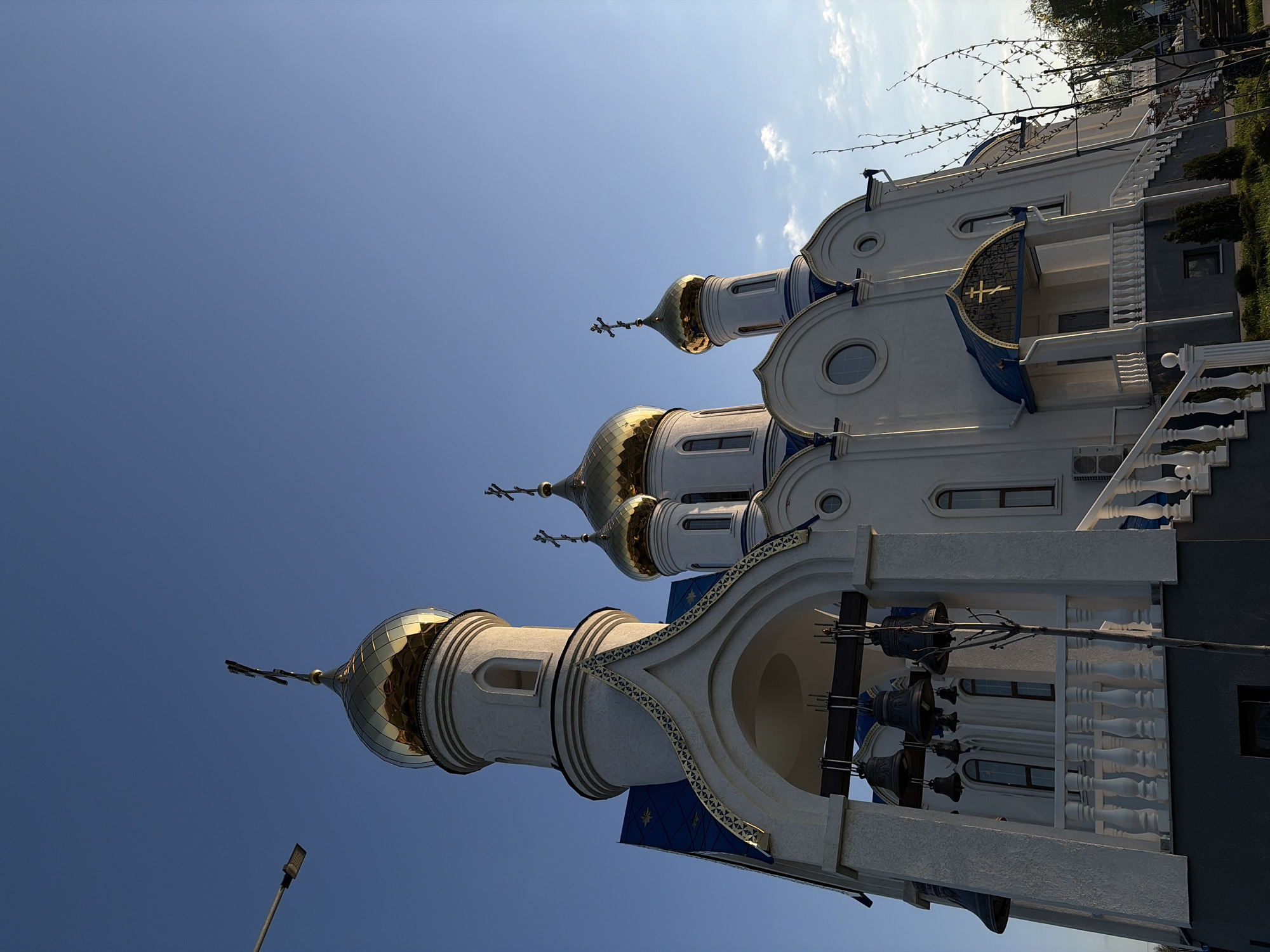 This image captures a low-angle, upward view of an ornate Orthodox church, located in Novopokrovskaya, Russia. The church is predominantly white with striking golden onion domes, each topped with a cross, set against a vast, clear blue sky dotted with a few wispy clouds.
The architectural style features intricate details, including arched windows and decorative blue elements along the cornices and beneath the domes. A prominent bell tower, visible in the foreground, houses several large, dark-colored bells. White balustrades adorn the balconies and staircases, adding to the building's grandeur. The bright sunlight illuminates the golden domes, making them gleam, and casts subtle shadows that highlight the church's intricate textures and three-dimensional form.
A street light is partially visible in the lower left, and some sparse tree branches appear in the upper right against the sky. The time of day is clearly bright and sunny. There are no people or specific activities taking place, with the focus solely on the impressive and serene architecture of the religious structure. No visible text is present in the image.