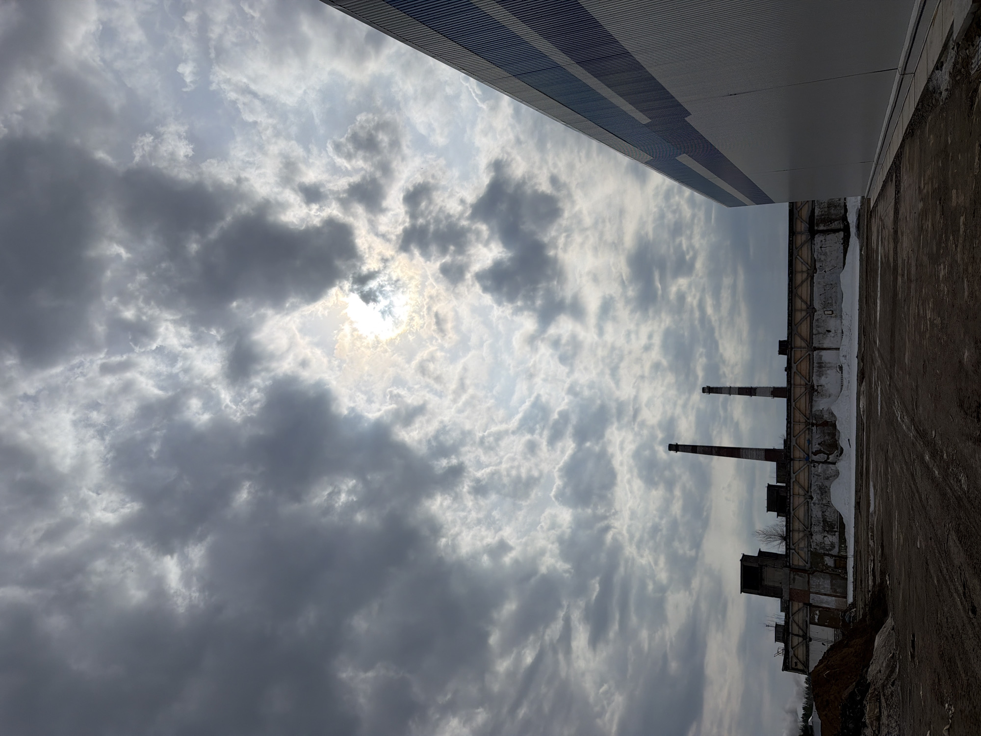 The photograph depicts an industrial landscape taken in Chaikovsky, Russia. The sky is filled with a thick blanket of grey and white clouds obscuring the sun. The lower portion of the image features a deteriorating industrial building with three prominent smokestacks rising from the structure. Adjacent to the building is a dirt or paved area, possibly a road or yard, with visible tracks and debris. Part of the roof of an adjacent large metal structure is visible on the upper edge.