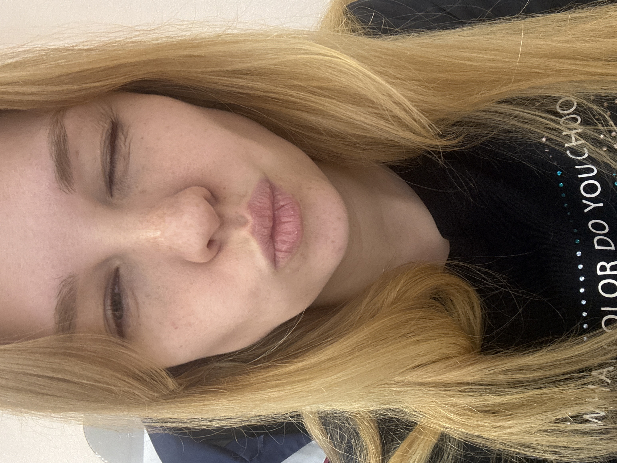 A close-up, rotated portrait of a fair-skinned individual with blonde hair, eyes closed. The person is wearing a black shirt with white text that reads "COLOR DO YOU CHOO". The location is not discernible from the image.
