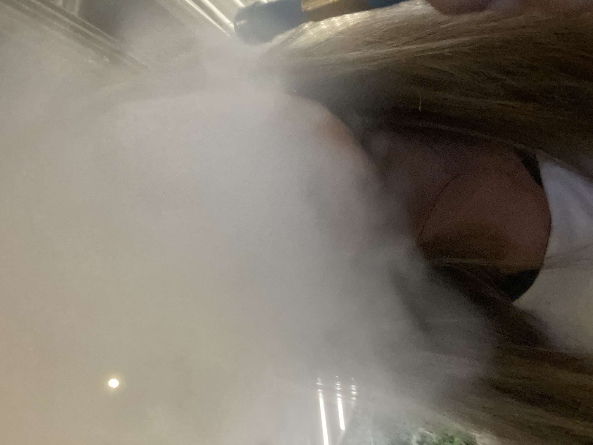 In an indoor setting, the photograph captures a close-up of a person's head and shoulders, obscured by a large plume of white vapor. The person has long, light brown hair that is visible on the sides of their head and falls down their shoulders. They are wearing a white garment, possibly a t-shirt, which is partially visible. To the right of the frame, a blue object with a golden tip is partially visible, held in the person's hand, suggesting the vapor is exhaled from a vaping device. The background is indistinct due to the vapor, but some horizontal lines and a bright light source are faintly discernible, possibly indicating an overhead light or architectural feature.