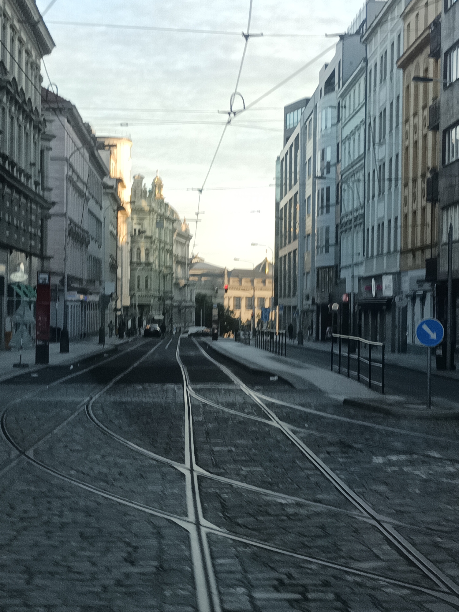 The image depicts a view of a street in Prague, Czechia, with tram tracks in the foreground. The perspective is from a low angle, looking down the street lined with multi-story buildings on both sides. The buildings feature classical architecture with detailed facades and numerous windows. In the mid-ground, a black car is visible driving away from the camera on the tram tracks. On the right side of the street, there is a blue circular sign with a white arrow pointing left. Overhead, tram wires are strung across the sky, indicating active tram infrastructure. The cobblestone street and the architectural style suggest an older European city, consistent with Prague. The lighting appears to be natural, possibly early morning or late afternoon, with soft sunlight illuminating parts of the buildings. The scene is devoid of any discernible human activity besides the driver of the car.