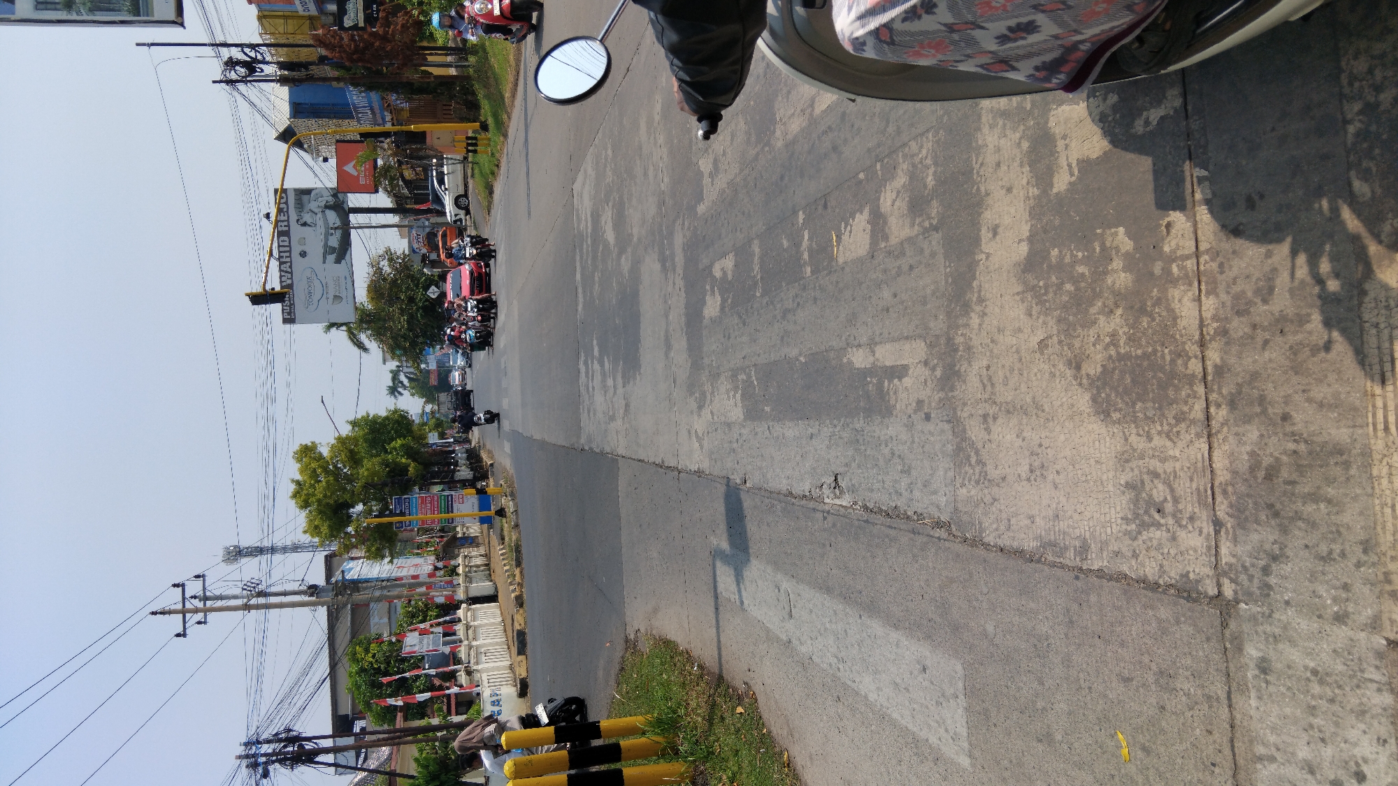 The image shows a street scene in Kauman, Indonesia, on a bright, clear day. The foreground is dominated by the road surface, with a pedestrian crossing marked by faded white stripes. To the left, a sidewalk with some grass and yellow and black striped poles is visible. In the middle ground, traffic is moving in both directions. Several motorcycles and a few cars are on the road. A prominent billboard advertising "PUSAT WAHID RESORT" is visible above the road in the distance, supported by a traffic light structure. On the right side of the image, a motorcycle's handlebars and mirror are in the foreground, suggesting the photo was taken from a moving motorcycle. Buildings line the sides of the road, with trees and power lines overhead. The overall impression is a typical urban street with moderate traffic and commercial signage.