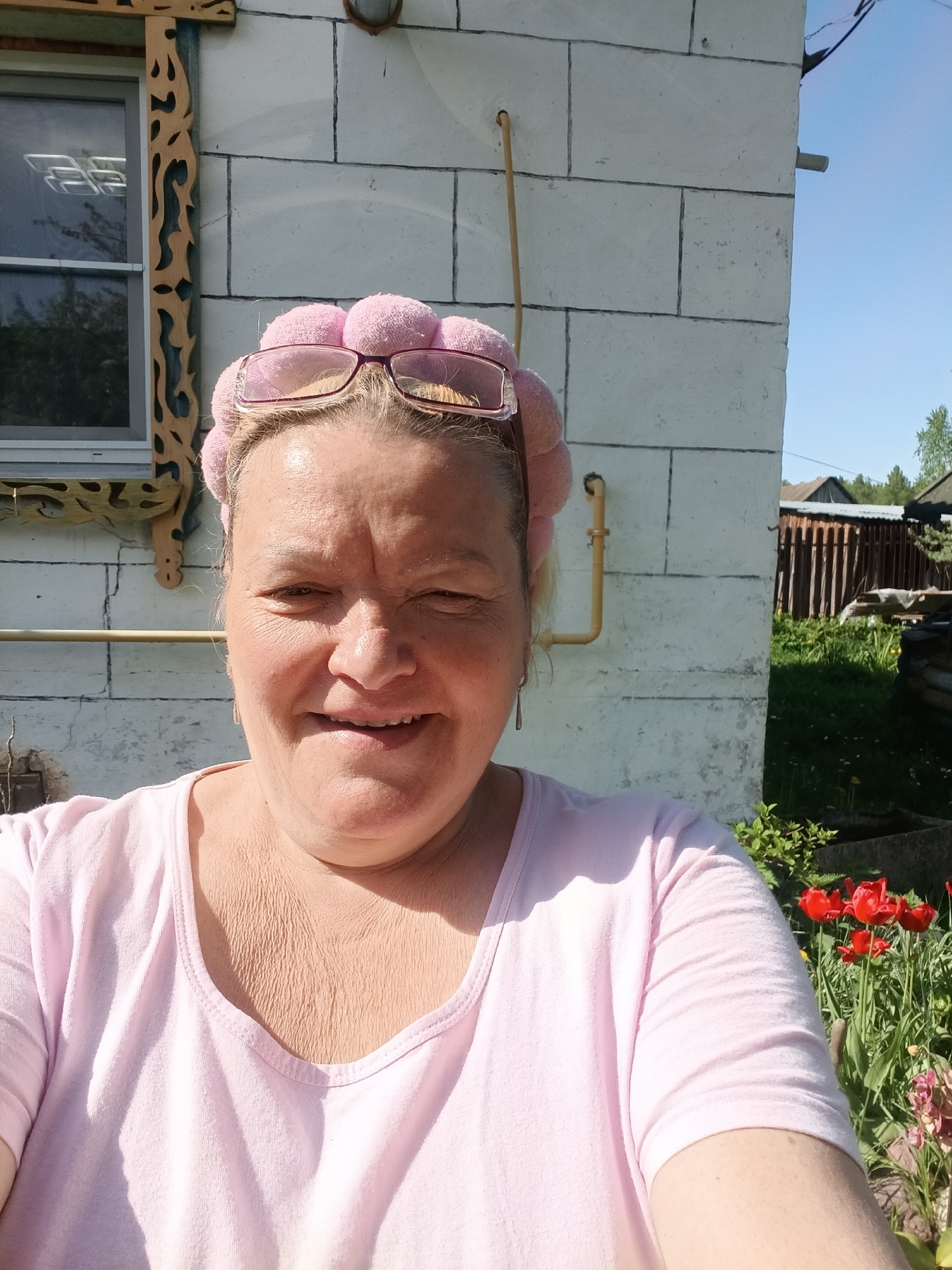 A medium shot captures a middle-aged woman in Красный Маяк, Russia. She is wearing a pink T-shirt, a pink headband, and pink-rimmed glasses on top of her head. The woman smiles at the camera. Behind her is a white brick building with a window framed in ornately carved wood trim. A yellow gas pipe runs vertically against the building. In the background, wooden fences, greenery, and red tulips are visible. The scene is illuminated by bright sunlight.