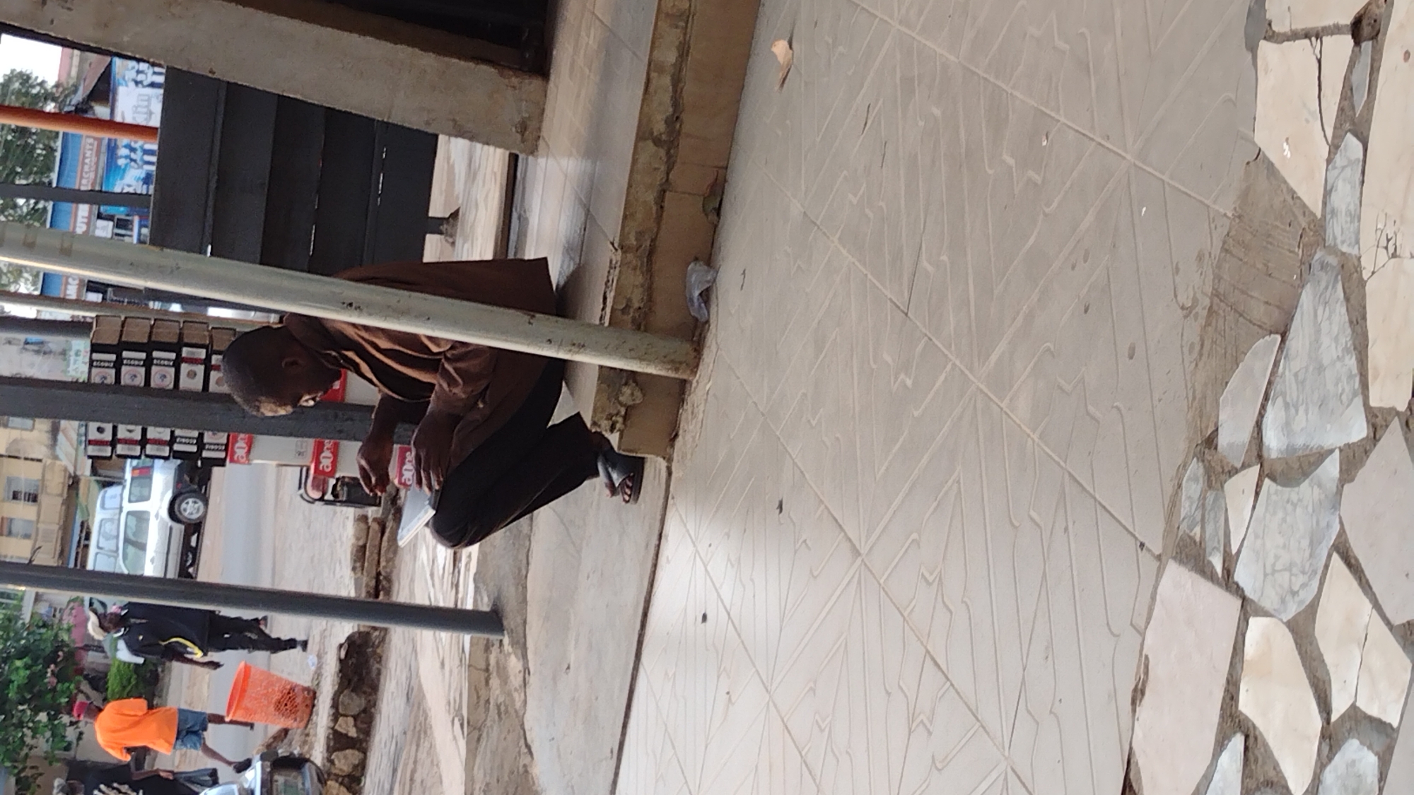 The image depicts a man sitting on the side of a tiled walkway near a building in Jos, Nigeria. He is wearing a brown shirt and dark pants and appears to be focused on something in his hands. A display of small items is visible behind him. In the background, a street scene includes a white van and pedestrians. The walkway is composed of square tiles and an area of irregularly shaped white tiles.