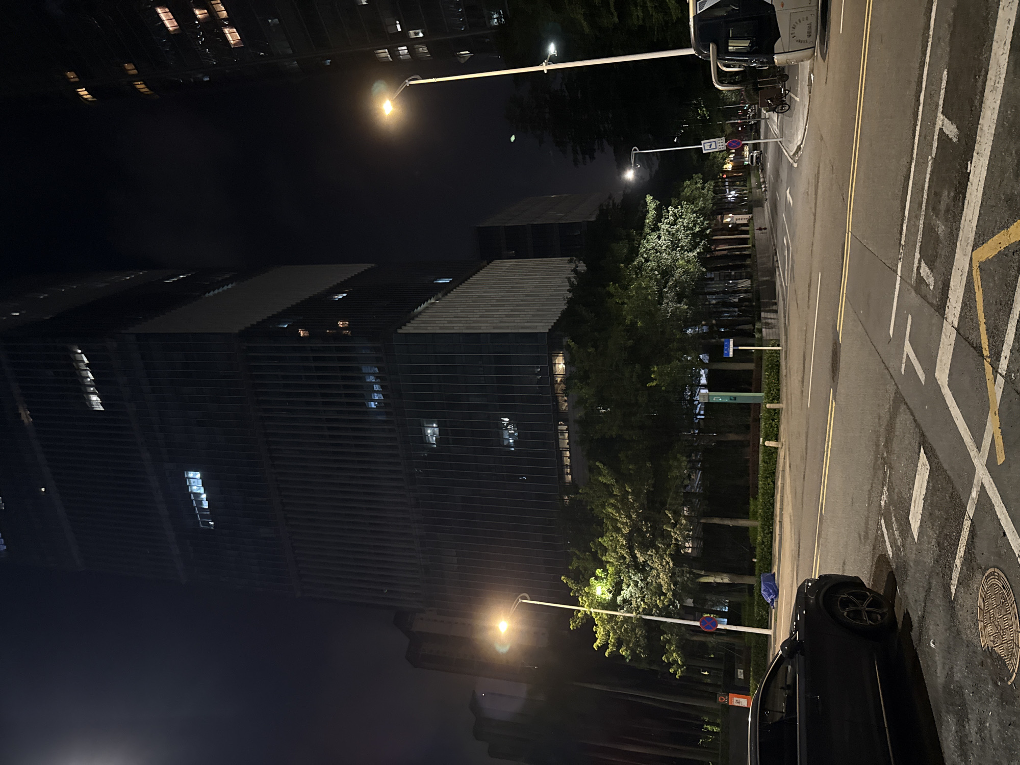 The image depicts a nighttime scene in a city, likely Haizhu District, China. Tall, modern buildings with illuminated windows dominate the left side of the frame, suggesting an urban environment. The street, marked with white and yellow lines and a yellow box junction, is wet, indicating recent rain or damp conditions.
A dark-colored car is parked on the right side of the road, its rear wheel clearly visible. In the background, trees with lush green foliage are illuminated by streetlights, creating a contrast between the natural elements and the artificial city lights. Streetlights are also visible along the road, casting a warm glow on the wet pavement.
There are several signs visible, including a blue rectangular sign with white text and a circular red sign with a blue background and a white horizontal bar, which appears to be a prohibition sign. A bus is parked on the left side of the street, with its front facing away from the camera. The overall atmosphere is one of quiet urban nightlife, with the city lights reflecting on the damp surfaces. The time of day is clearly night, and the weather appears to be clear despite the wet roads.