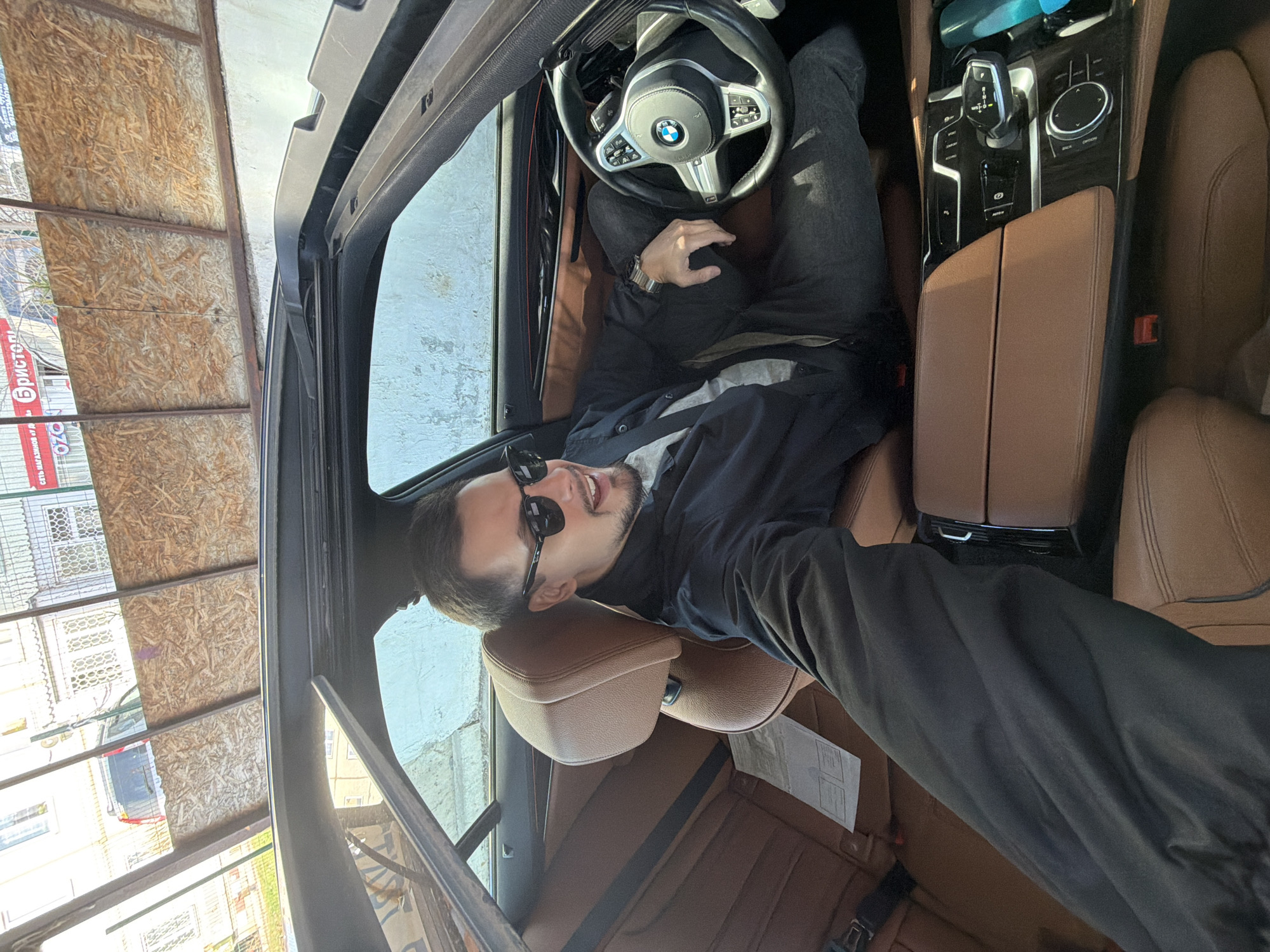 The image captures a man inside a car, appearing to be taking a selfie or being photographed from the rear seat. The man, with a beard and dark hair, is positioned on the front passenger seat, leaning back with his head resting on the brown leather headrest. He is smiling widely, revealing his teeth, and wearing dark sunglasses, a dark jacket over a grey t-shirt, and dark trousers. A metallic watch is visible on his left wrist.
The car's interior features brown leather seats and a modern dashboard. The steering wheel, partially visible in front of where the driver would sit, displays the BMW logo and an 'M' logo, indicating a BMW vehicle. The center console is equipped with a gear selector, an iDrive controller, and buttons for driving modes such as "SPORT", "COMFORT", and "ECO PRO". A white document is lying on the rear passenger seat.
Through the car's windows, exterior details are visible under bright daylight. On the left side (driver's side view), there's a structure composed of wooden panels, possibly OSB, arranged horizontally. Further in the background, a building with white walls and intricate window grilles can be seen. On a red sign visible outside, Cyrillic text elements like "БРИСТОЛ" (Bristol) and "OZO" (likely Ozon) are discernible. The man's relaxed posture and smile suggest a casual and content mood. The setting appears to be urban or semi-urban.