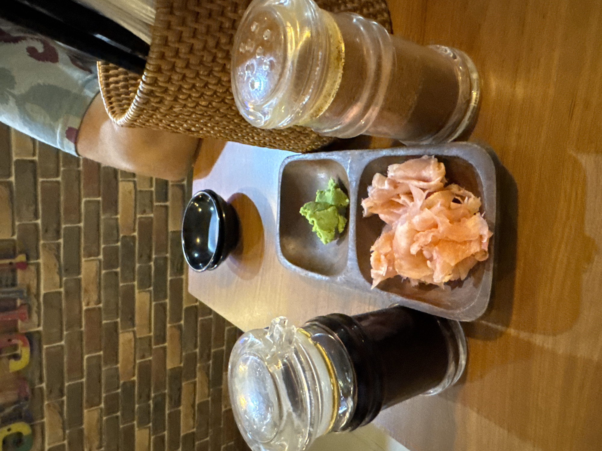 An overhead, slightly angled shot captures a dining table setting, likely in an indoor restaurant environment. Central to the image is a rectangular wooden or dark ceramic tray with three compartments. One compartment contains a small, empty black ceramic bowl, presumably for dipping. Adjacent to it, a vibrant green paste, identifiable as wasabi, is present. The third compartment holds a pile of thinly sliced pink pickled ginger (gari).
Two glass condiment shakers are also on the table. One, located in the upper right, contains a light brown granular substance, possibly a spice or seasoning. The other, positioned in the lower left, is a bottle with a dark liquid, clearly soy sauce. A woven wicker basket, partially visible on the left side of the frame, holds several black chopsticks.
Part of a person's left forearm and shoulder, wearing a light blue or grey short-sleeved top with a subtle, faded pattern, can be seen on the far left. The table surface is light brown wood. In the background, a wall features rectangular brick-like tiles in various shades of brown, grey, and tan. This setup strongly suggests a meal of sushi or sashimi is either about to be consumed or has just begun. On the lower left part of the brick wall, faint, colorful, stylized letters are visible, but too blurry and incomplete to be read definitively.