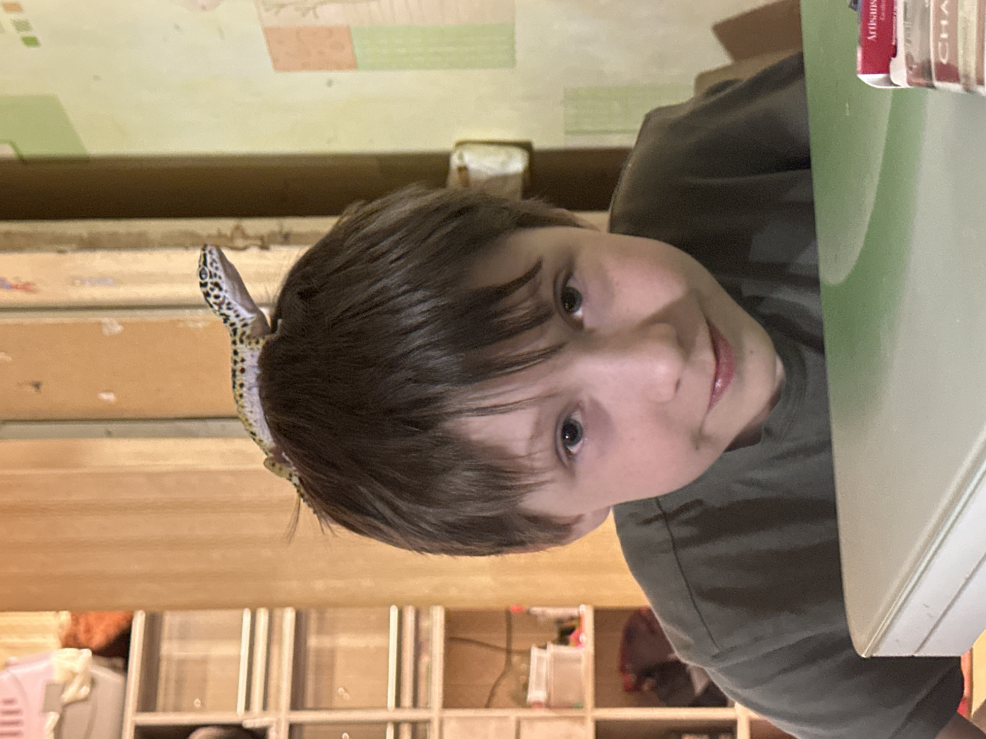 A young person with short brown hair is looking at the camera. A gecko is perched on top of their head. They are sitting at a table with books visible on the right edge. The background includes a wall with patterned wallpaper and a shelf with various items. There is no indication of the location being Moscow, Russia.