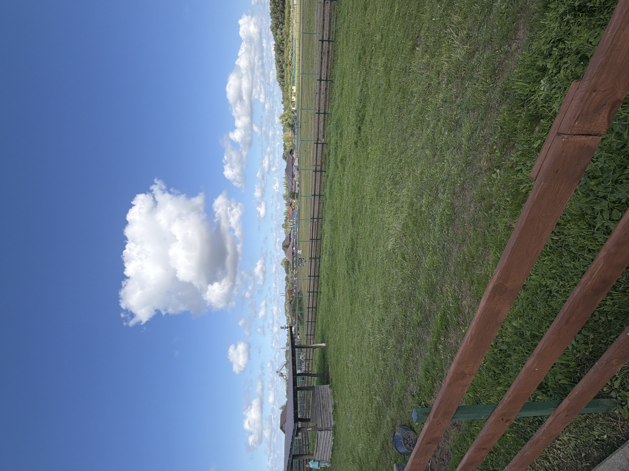 This is a photograph of a rural outdoor scene. In the foreground, there is a wooden fence with brown planks. Behind the fence is a large, grassy field. To the left, there is a wooden shelter with a dark roof, and a person wearing a light blue shirt is standing near it. In the middle ground, a metal fence runs horizontally across the image, separating the grassy field from another area. Beyond this fence, there are several buildings, including houses with red roofs, and some trees. The background consists of a distant treeline and a bright blue sky with scattered white clouds. The image appears to be taken in Konstantinovka, Russia.
