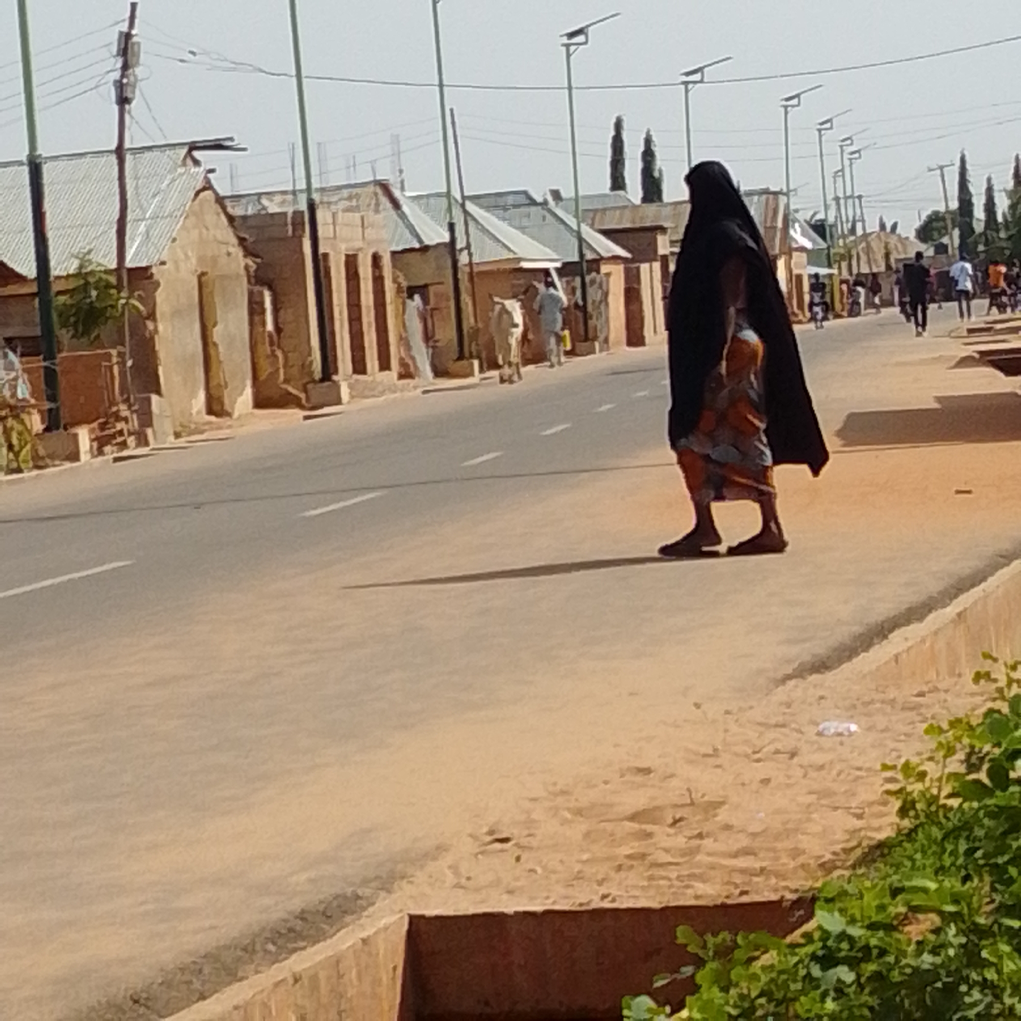 This image captures a street scene in Gombe, Nigeria. In the foreground, a person wearing a black hijab and a colorful patterned skirt walks along a dirt path next to a paved road. The path has a drainage ditch on its outer edge, with some green foliage growing nearby. The road itself has white lines marking its lanes.
Along the side of the road, there are several low-lying buildings with corrugated metal roofs and rough, unfinished walls. Some of these buildings appear to be shops or market stalls. In the background, more buildings and trees are visible, as well as other people walking and some motorcycles. The sky is overcast, and the overall atmosphere suggests a typical day in a bustling town or city.