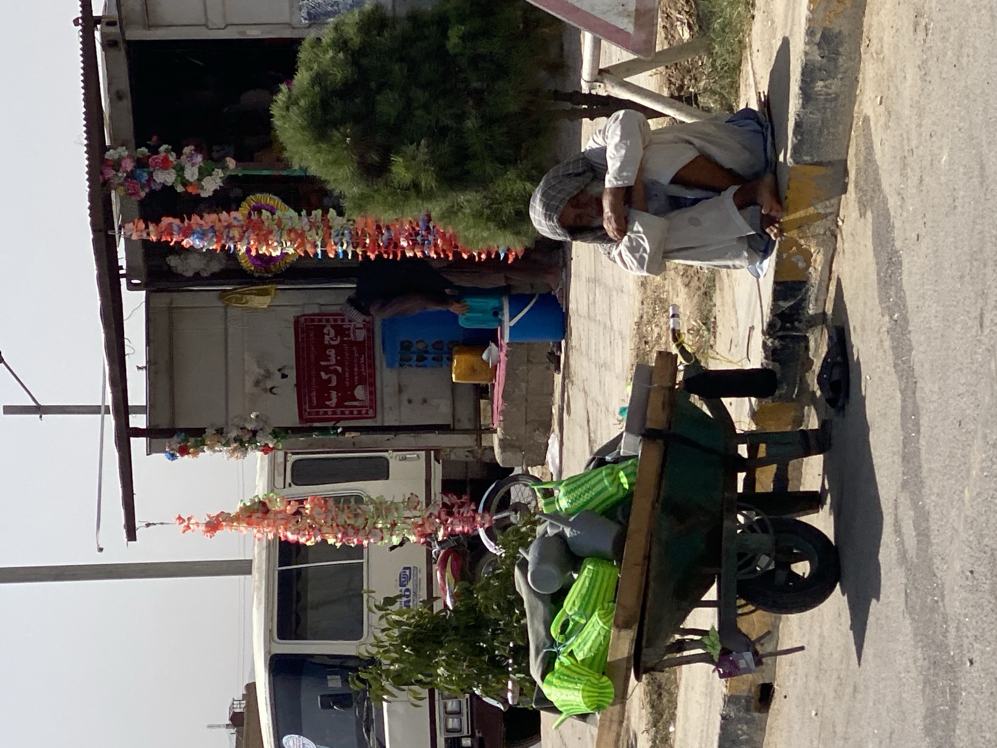 The image depicts a street scene in Kandahar, Afghanistan. In the foreground, a man sits on a curb, his legs crossed, wearing a checkered headscarf and a light-colored tunic. His eyes are closed and his head is bowed. To his left, a green wheelbarrow is laden with various items, including several bright green watering cans and some gray objects. The wheelbarrow is on a paved road next to a raised curb.
Behind the wheelbarrow, a white bus is partially visible. Hanging from an overhead structure, there are colorful floral garlands, suggesting a festive or decorative element in the environment. Further back, a small shop or stall is visible, with a person inside and some containers on the ground. The overall environment appears to be a roadside, possibly a marketplace or a busy intersection, with a clear sky overhead. The ground is a mix of paved road and dirt or gravel.