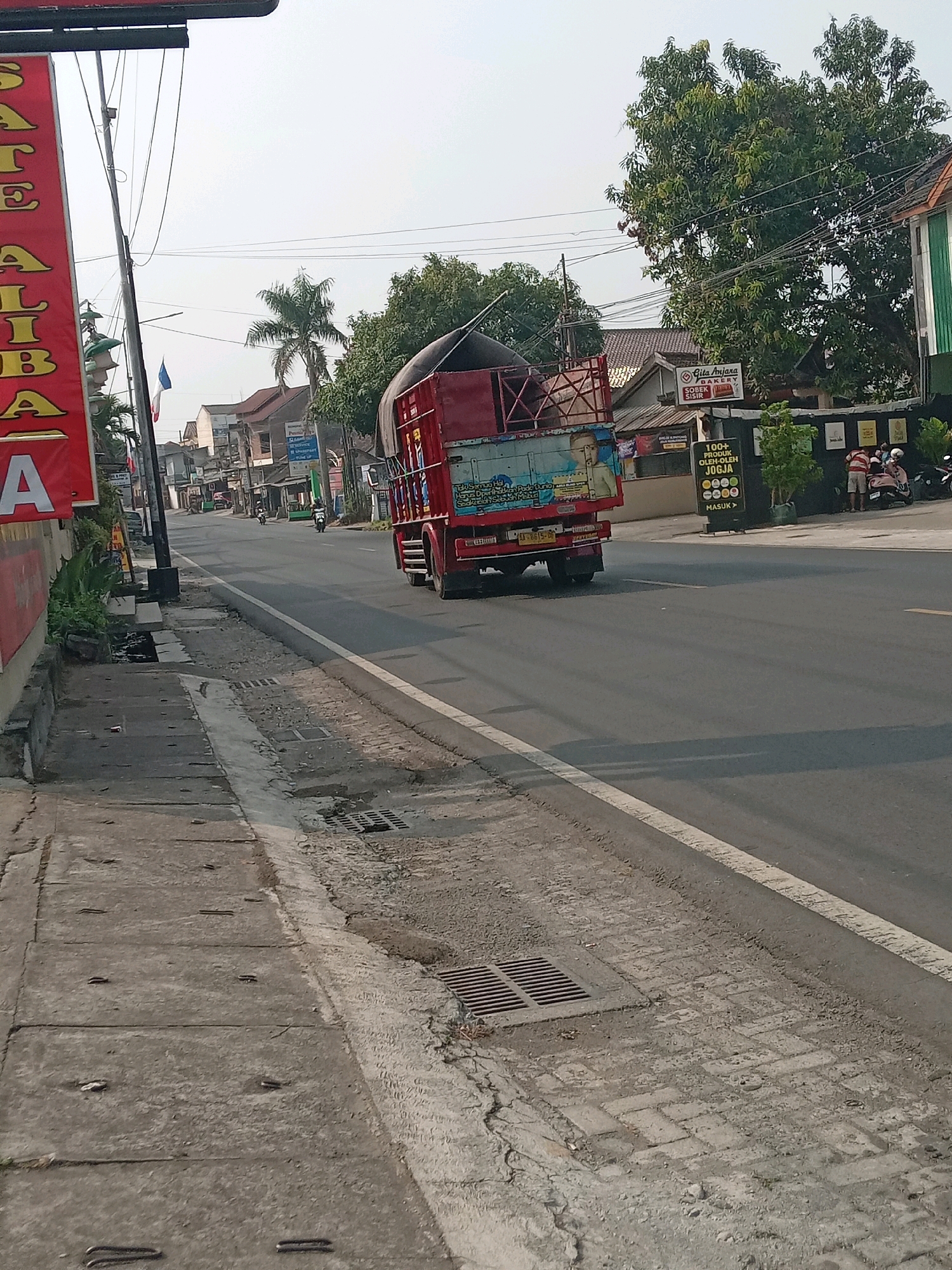 The image depicts a street scene in Banguntapan, Indonesia, likely on a main road given the traffic and commercial establishments. A red truck with a white tarp-covered load is a prominent subject, moving away from the viewer. The truck's side features a colorful mural with figures and text. On the left side of the road, a red banner with large, vertical yellow text, partially obscured by a pole, is visible, suggesting a business advertisement. Further down the street, several buildings line both sides, indicating a commercial or mixed-use area. Signs are present, including one for "Gita Anjana BAKERY" and another displaying "100+ PRODUK OLEH-OLEH JOGJA" with circular graphics, pointing to a food or souvenir shop. A white and green building is visible on the right. Several people are seen near the buildings, some standing and some on motorcycles, suggesting everyday street activity. A palm tree and other leafy trees add to the tropical environment. The road is paved, with a distinct white line marking the lane. A concrete sidewalk runs along the left side of the road, featuring drainage grates and some wear and tear. The sky is overcast, suggesting daytime. The overall impression is of a typical bustling street in a Indonesian town.