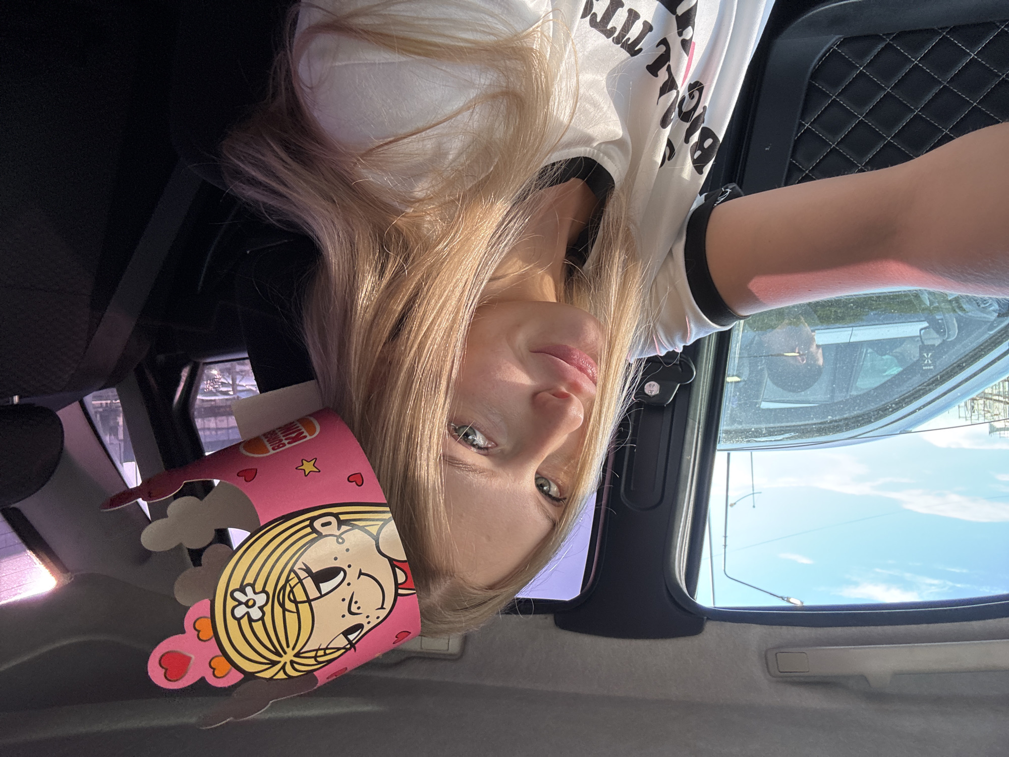 In an interior shot of a car, a young woman with blonde hair and light green eyes is seen wearing a pink party hat with a cartoon character. She is looking towards the camera and smiling. The woman is wearing a white t-shirt with black trim and text that is partially obscured. Her left arm is visible, with a pink hue on her skin, possibly from the sun or the lighting. The background shows the car's interior, including a black seatbelt and a car door with quilted detailing. Through the car window, other vehicles and a cloudy sky are visible. The scene takes place during the daytime. There is no specific identifiable landmark or context to place this image in the city of Perm, Russia.
