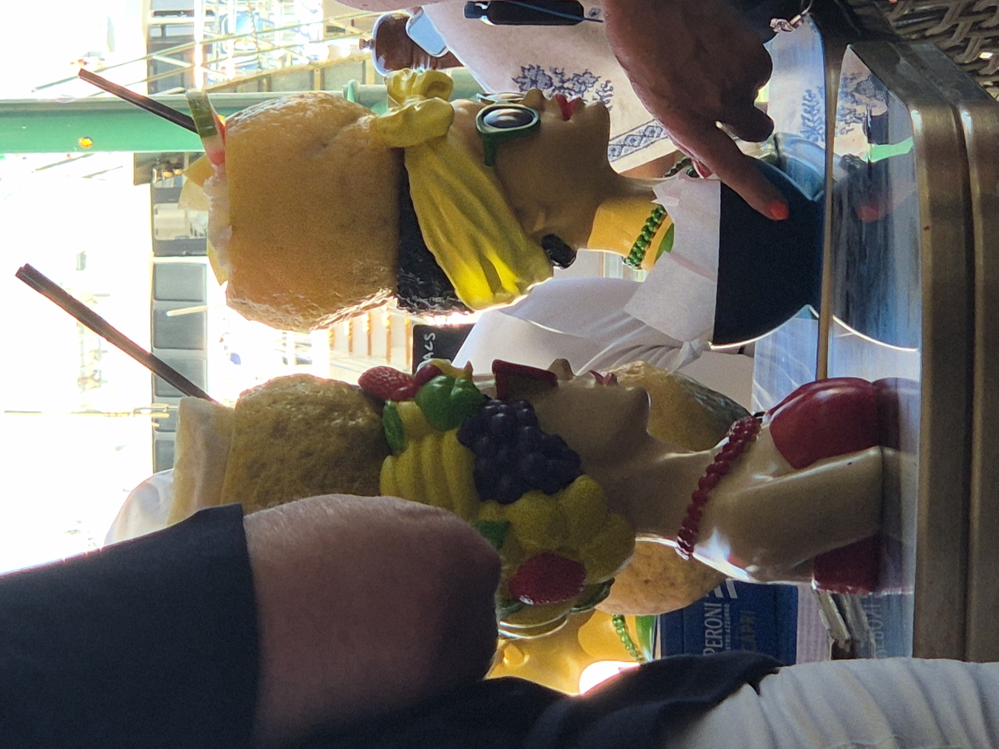 In an outdoor setting, likely a cafe with a waterfront view, two decorative busts of women are prominently displayed on a table. The bust on the left has a headpiece resembling a crown of colorful fruits, including grapes and strawberries, and is adorned with a red beaded necklace and a red garment. The bust on the right wears a yellow headscarf and green sunglasses, with a yellow collar. Both busts have exaggerated features and appear to be made of resin or plastic. Behind them, a person's arm and hand, with orange nail polish, are visible, seemingly interacting with the scene. In the background, a Peroni Capri beer box is visible, and parts of a boat and railing can be seen, suggesting the location is near a harbor or waterfront. The lighting is bright, indicating it is daytime.