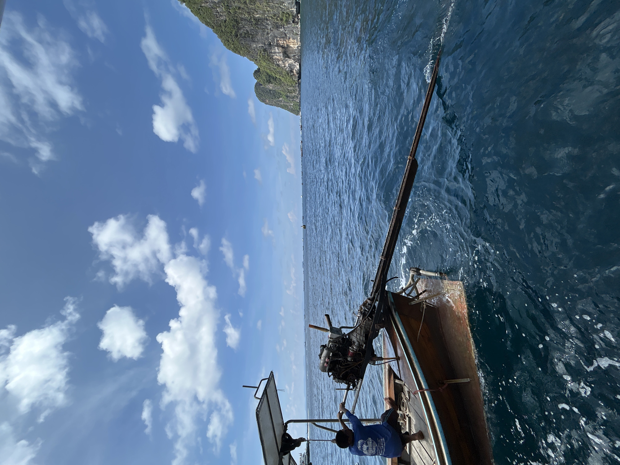 A longtail boat is in the foreground, its wooden hull visible as it cuts through deep blue water. A person wearing a blue t-shirt and shorts sits at the back of the boat, near a prominent engine with a long tail shaft extending into the water. The boat is moving, indicated by the wake behind it. In the background, a steep, tree-covered limestone cliff rises dramatically from the sea, characteristic of the landscape around Ao Nang, Thailand. The sky is a bright blue with scattered white clouds. Several other boats are visible on the horizon, suggesting a popular tourist area.