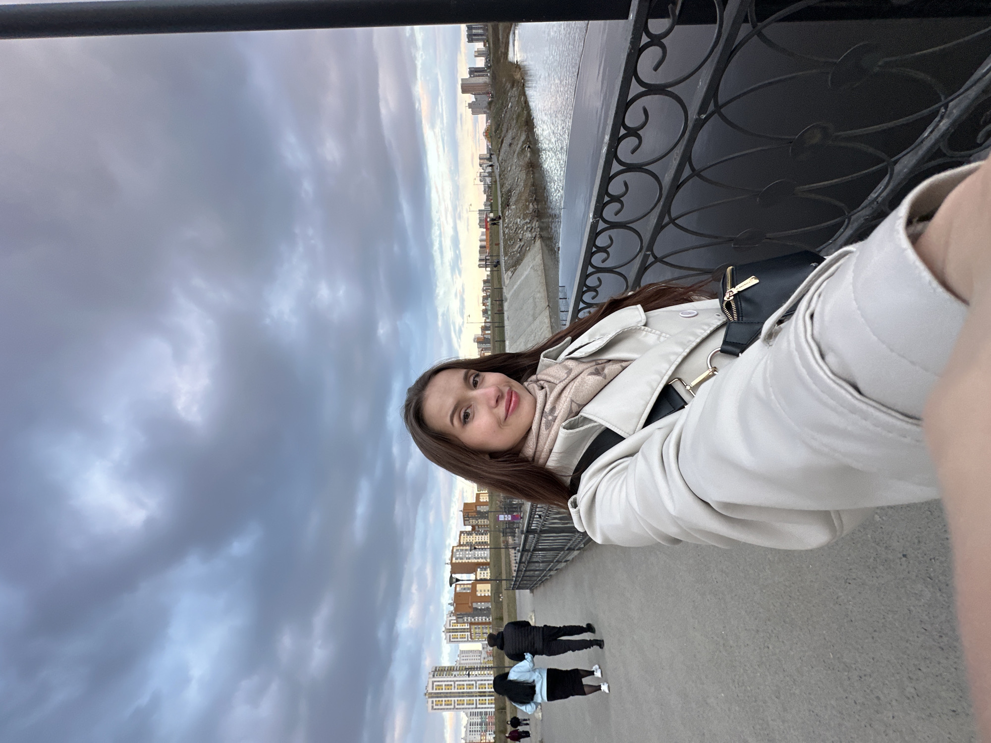 A photograph captures a young woman posing in front of a bridge with elaborate metal railings. The woman has long brown hair, fair skin, and is wearing a light-colored coat and a patterned scarf. Behind her, a riverside embankment stretches out, revealing the skyline of Yekaterinburg, Russia, with its tall buildings. On the pedestrian walkway below, several people are visible, including a couple walking together. The sky is overcast with dramatic, grey clouds.