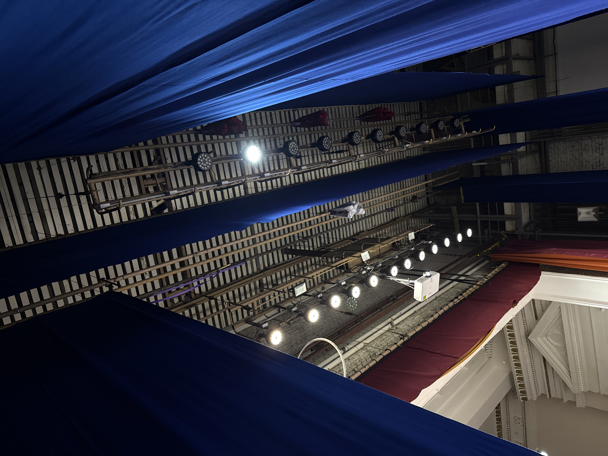 The image shows a view looking up at the ceiling of what appears to be a theater or event space. The ceiling is constructed with an exposed grid of beams, some of which have lighting fixtures attached. The fixtures are of varying types, including multi-LED spotlights and spherical bulb lights. Fabric drapes in varying shades of blue are suspended, creating a layered visual effect. Additionally, there are hints of other draped fabrics in maroon and orange. At the bottom right, architectural details suggest a more ornate part of the building, possibly a balcony or mezzanine level. The location (Perm, Russia) cannot be verified with certainty from the image alone, however, the architectural details do not contradict such a scenario.