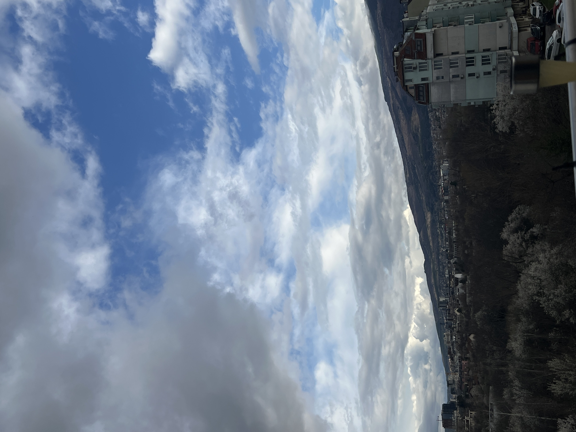 The image depicts a cityscape in Sofia, Bulgaria, juxtaposed against a dynamic sky. The sky is filled with varying types of clouds, ranging from light, wispy cirrus formations to darker, more ominous cumulonimbus clouds. Interspersed among these clouds are patches of clear blue sky.
In the foreground, there are buildings of light color, along with trees and cars. A mountain range is located near these buildings.