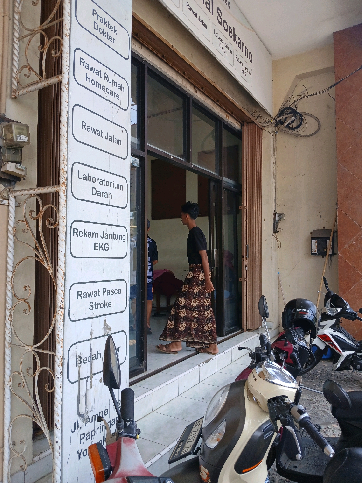 The image shows the exterior of a clinic or medical facility, likely named "Rumah Sakit Soekarno" based on the signage visible at the top. A vertical signboard on the left lists various services such as "Praktek Dokter" (Doctor's Practice), "Rawat Rumah Homecare" (Home Care), "Rawat Jalan" (Outpatient), "Laboratorium Darah" (Blood Laboratory), "Rekam Jantung EKG" (ECG Heart Record), "Rawat Pasca Stroke" (Post-Stroke Care), and "Bedah" (Surgery). The clinic appears to be situated in a commercial area, with motorcycles parked in front. The entrance features a glass door and a brown, corrugated metal gate. A young person wearing a black t-shirt and a patterned sarong is seen walking into the clinic. The environment suggests a typical urban setting in Depok, Indonesia, with visible electrical meters and tangled wiring on the wall.