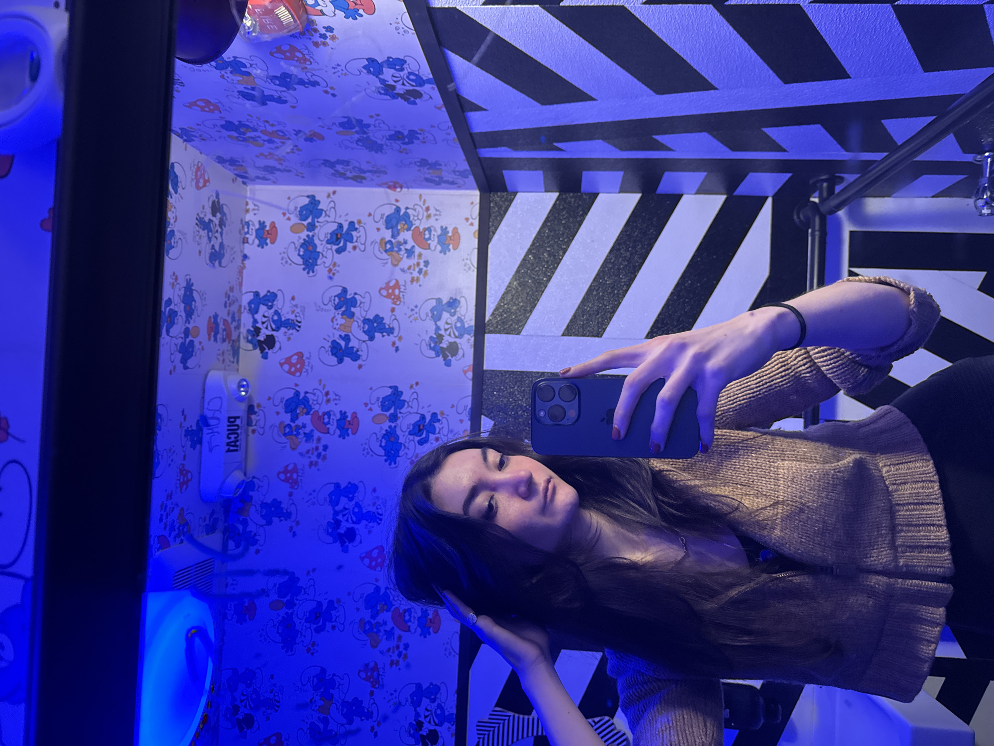 A selfie captures a young woman in a room with unique decor. She holds a black smartphone, her brown hair draped over her left shoulder. She is wearing a tan, knitted jacket. The room's walls feature wallpaper adorned with a cartoon smurf pattern, complemented by black and white striped designs. The illumination casts a blue hue. A toilet and sink are visible. While the city location is unknown, it is presumed to be in New York City, United States of America.