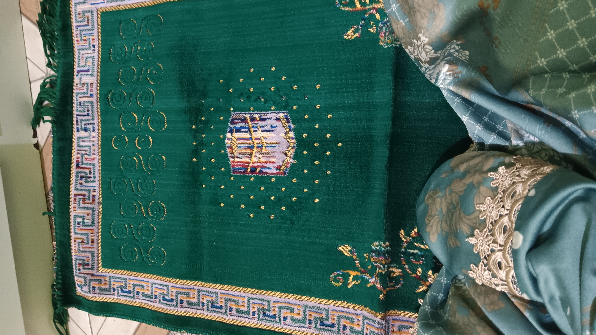 The image shows a green prayer rug with an embroidered depiction of the Kaaba at its center. The rug has intricate geometric patterns along its borders and is adorned with small golden dots surrounding the Kaaba image. In the foreground, a partially visible person is wearing a teal-colored garment with floral patterns and decorative lace trim on the sleeve. The prayer rug is laid out on a tiled floor, and a portion of a light-colored wall is visible in the background. The scene appears to be an indoor setting within a residential or religious space. The image does not provide information specific to Sonobekel, Indonesia.