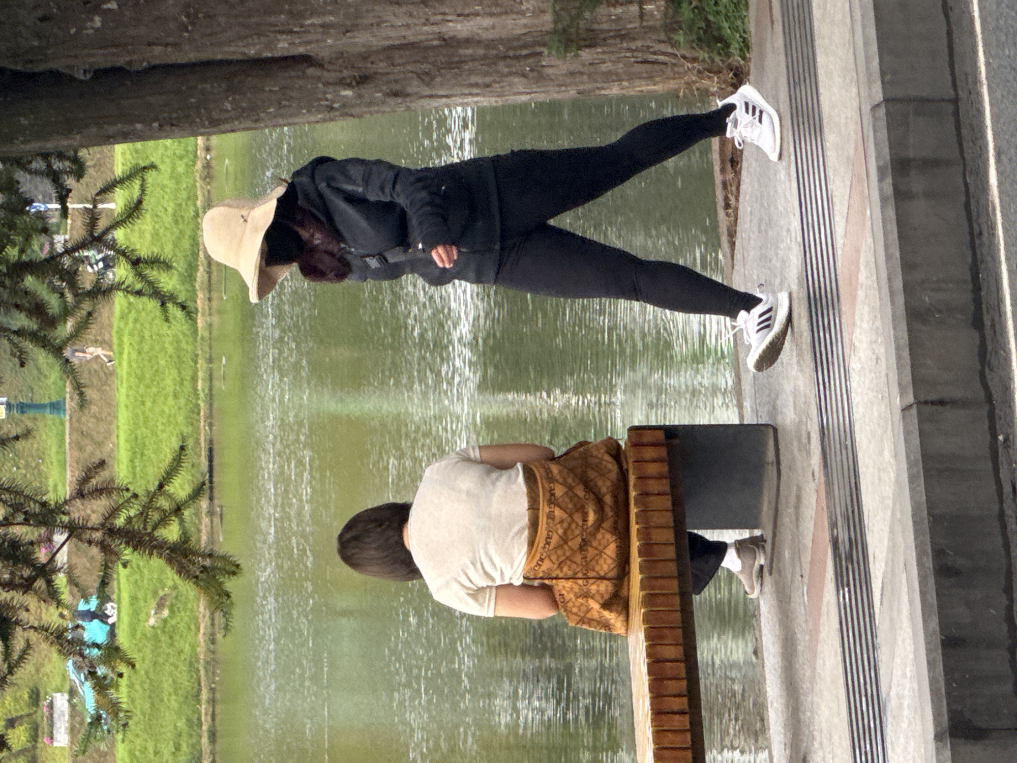 The image depicts two women near a body of water, likely Xuan Huong Lake in Da Lat, Vietnam, given the ambiance. The woman on the left, facing away from the camera, is seated on a wooden bench. She has dark, short hair and is wearing a light-colored t-shirt and a patterned, orange-brown skirt or sarong. The woman on the right is walking and facing towards the left side of the frame. She is wearing a dark hooded jacket, black leggings, and white athletic shoes with black stripes. She also has a light-colored wide-brimmed hat and a dark mask covering her face. A dark bag is slung across her body. The environment features a tree trunk on the right side of the image and a body of water with gentle ripples in the background. The ground in the foreground consists of a paved walkway with textured lines and a concrete curb. The overall scene suggests a casual outdoor setting.