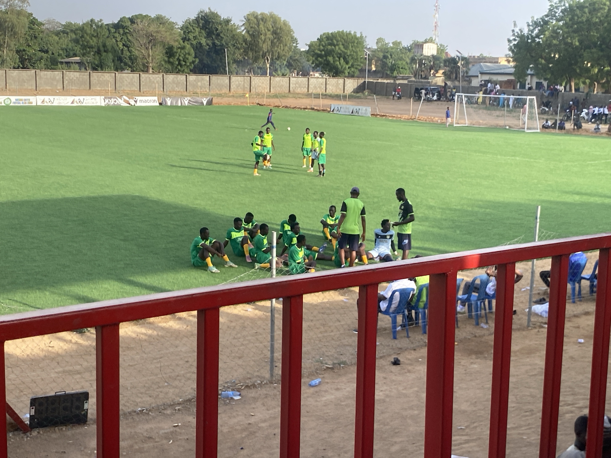 The image depicts a football field in Tudun Wada, Nigeria. A group of players in green and yellow uniforms are on the field, some sitting and others standing. A coach or manager is interacting with them. In the background, more players are practicing, and spectators are visible near the goal. A red railing is in the foreground, partially obscuring the view of the field. Debris is scattered on the ground behind the railing. Trees and a building surround the field, adding to the environment. The sky is overcast.