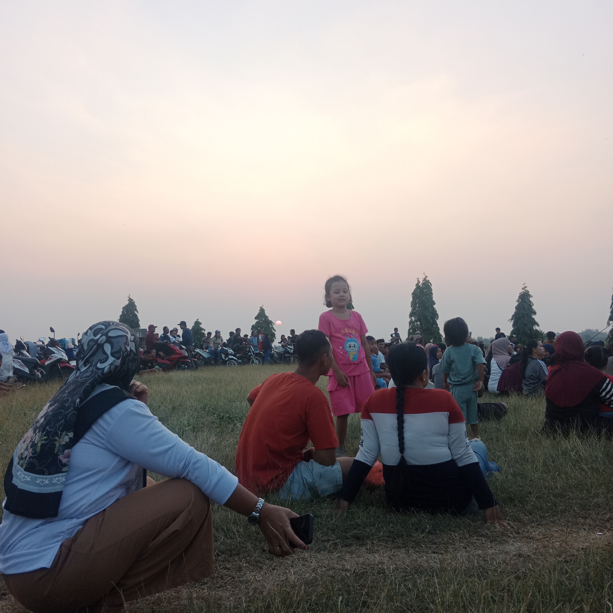 This image captures a gathering of people on a grassy field in Tegowanu Wetan, Indonesia, during what appears to be a sunset or twilight. The sky is a soft gradient of pale pinks and oranges, suggesting the end of the day. In the foreground, several individuals are seated or crouching on the grass. A woman in the lower left is wearing a blue long-sleeved shirt, brown pants, and a patterned headscarf. To her right, a person in an orange shirt and light blue shorts is seated. Another individual in the mid-right is wearing a red, white, and black horizontally striped shirt with a long black braid. In the background, a crowd of people and numerous motorcycles are visible, indicating a public event or casual meeting. Two tall, slender trees stand out against the sky in the middle ground. A young girl in a bright pink t-shirt and shorts is standing and looking towards the right side of the frame. The overall scene is one of community gathering in an outdoor setting.