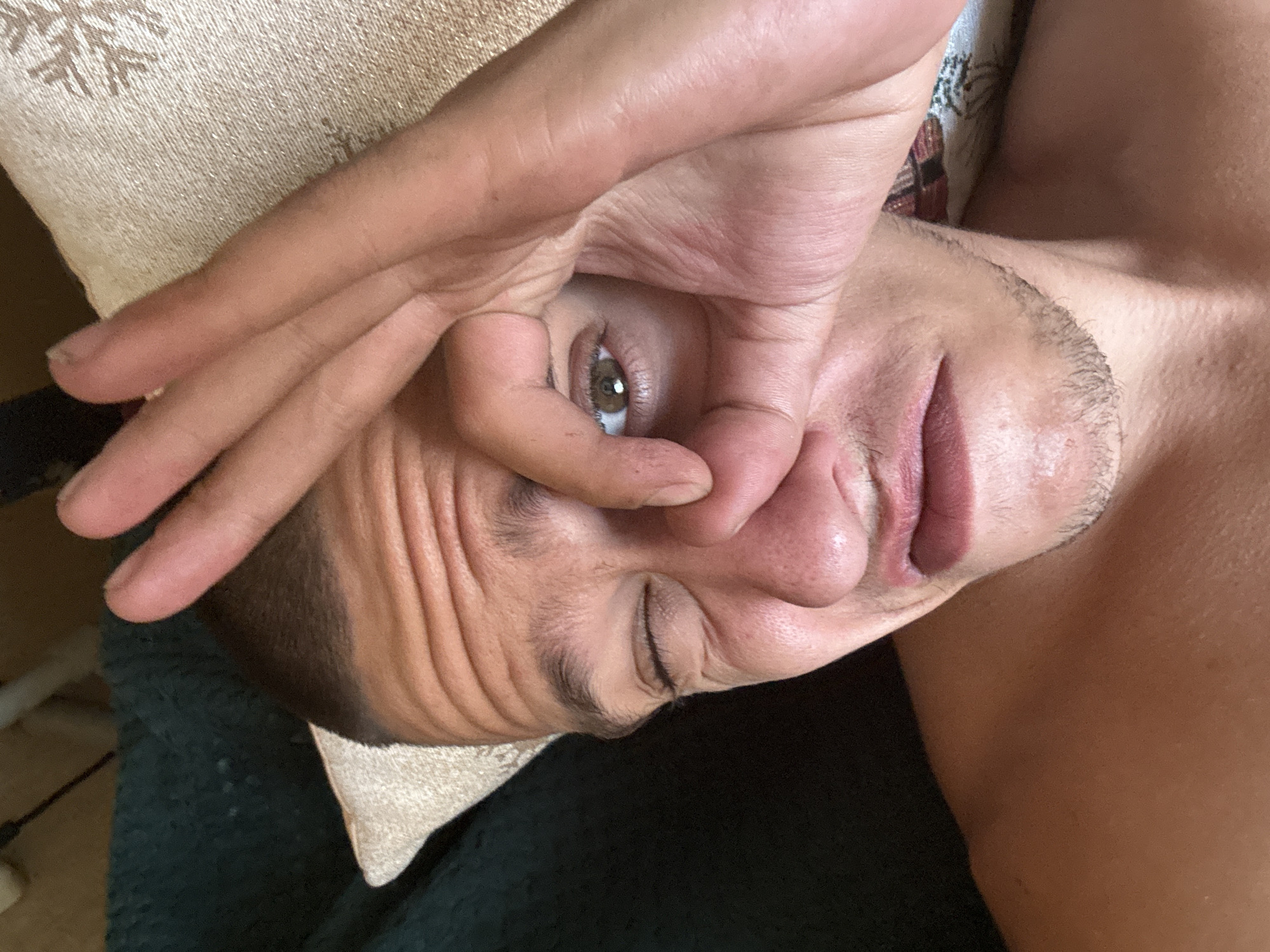 A close-up, selfie-style photograph shows a person's face from a slightly elevated angle. The person, appearing to be male, has a light brown complexion, short dark hair, and is shirtless. He is looking at the camera with his right eye open and his left eye squinted shut. His right hand is raised, with his thumb and index finger forming a circle around his right eye, while his other fingers are extended upwards. His mouth is slightly open in a grimace. The background consists of a dark green textured surface on the left, and a light-colored textured cushion with a snowflake pattern on the right. The overall lighting suggests an indoor setting. No specific location in Sofia, Bulgaria is identifiable from the image.