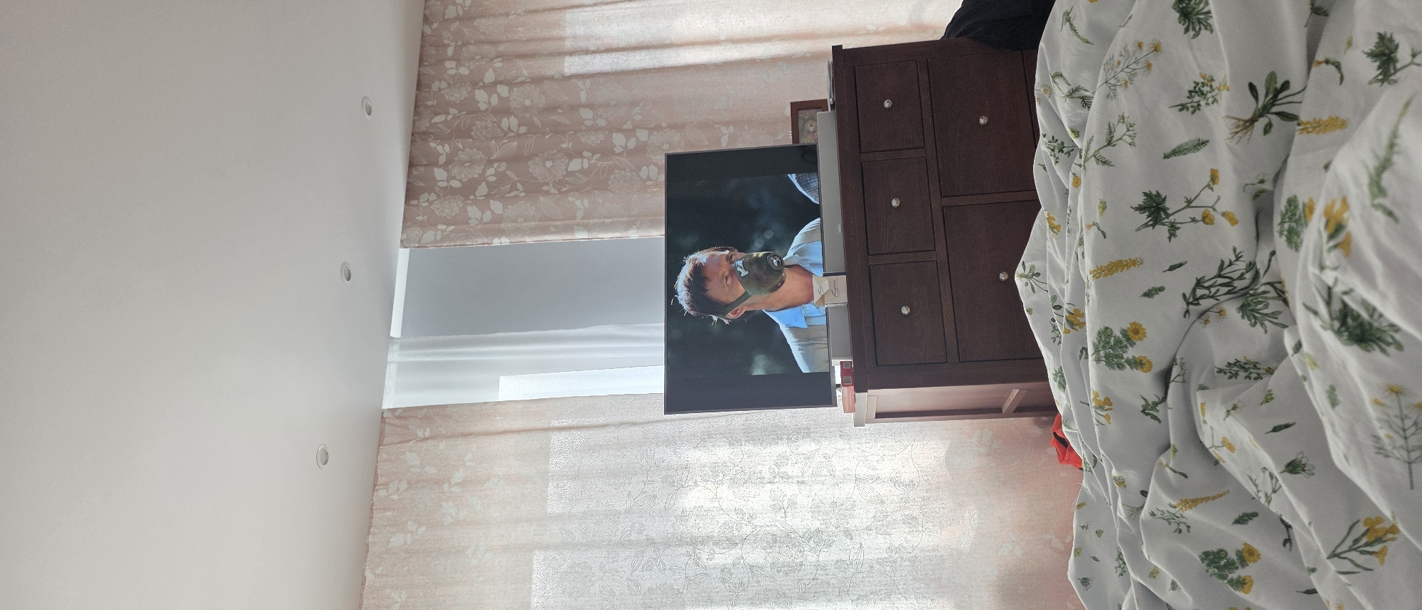 A man wearing a gas mask is visible on a television screen in a bedroom. The television is placed on top of a dark wood dresser with several drawers. The room has light-colored curtains with a floral pattern, and a white comforter with a botanical print is in the foreground. The ceiling has recessed lighting.