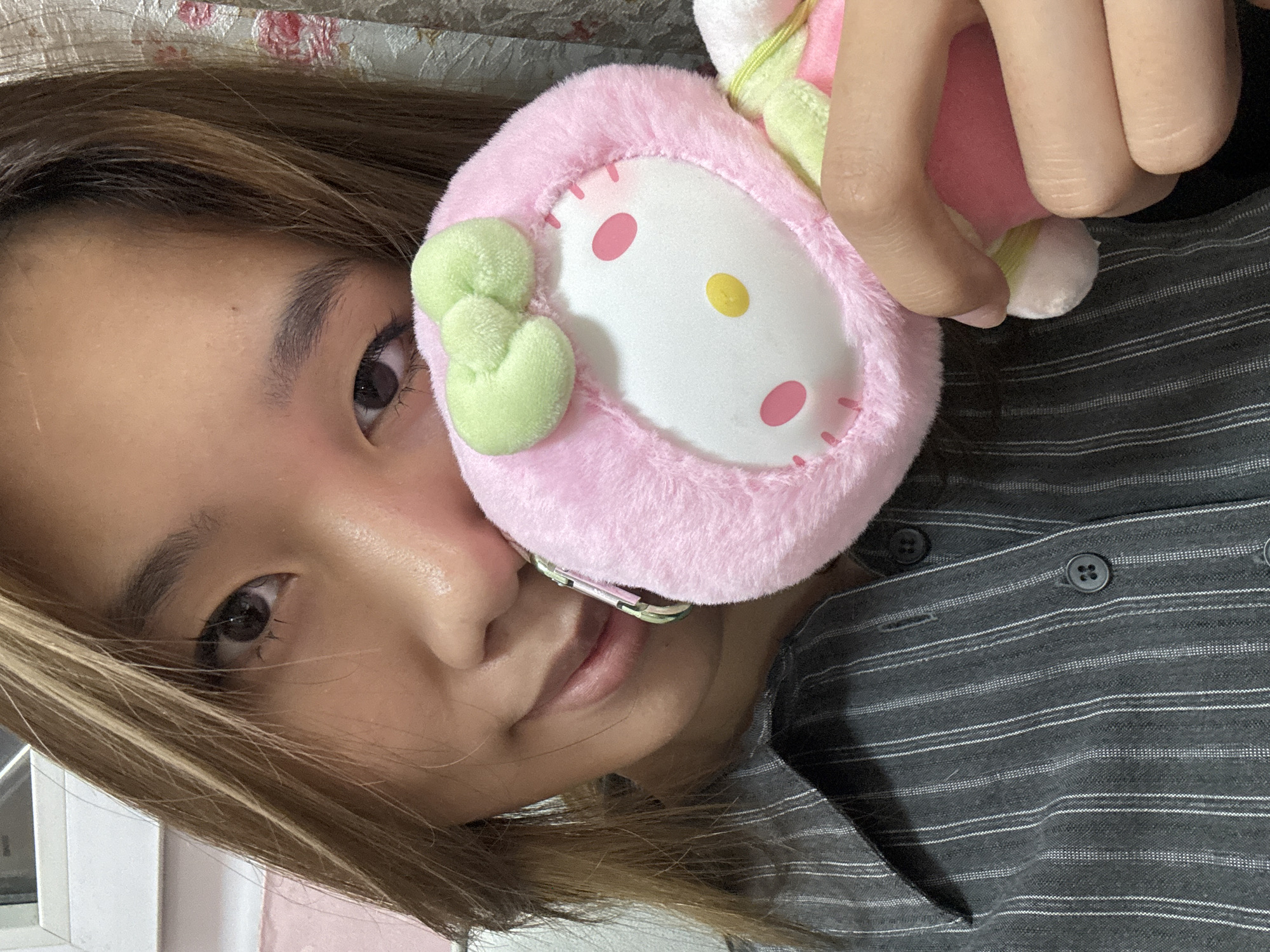 The image is a close-up, tilted portrait of a young person, likely female, with light brown hair, holding a plush toy. Her left eye is visible, looking directly at the viewer, and her hair frames the side of her face. A metallic dental brace is subtly visible on her lower teeth. She is wearing a dark gray or black button-up shirt with thin white horizontal stripes and visible buttons.
She is holding a pink, fluffy plush toy close to her face, partially obscuring her mouth and nose. The plush toy features a white oval face with two pink circular eyes, a yellow oval nose, and pink dash marks around its perimeter, typical of a Sanrio character like My Melody. A light green bow is affixed to the toy's left side. The person's fingers are visible gripping the toy near the top.
The background is indistinct but suggests an indoor setting, with a patterned fabric visible behind her head and a plain light surface on the far left. The lighting appears bright and natural, indicating it is likely daytime. There is no visible text in the image. The scene captures a casual, intimate moment. This image is set in Shapingba District, China.