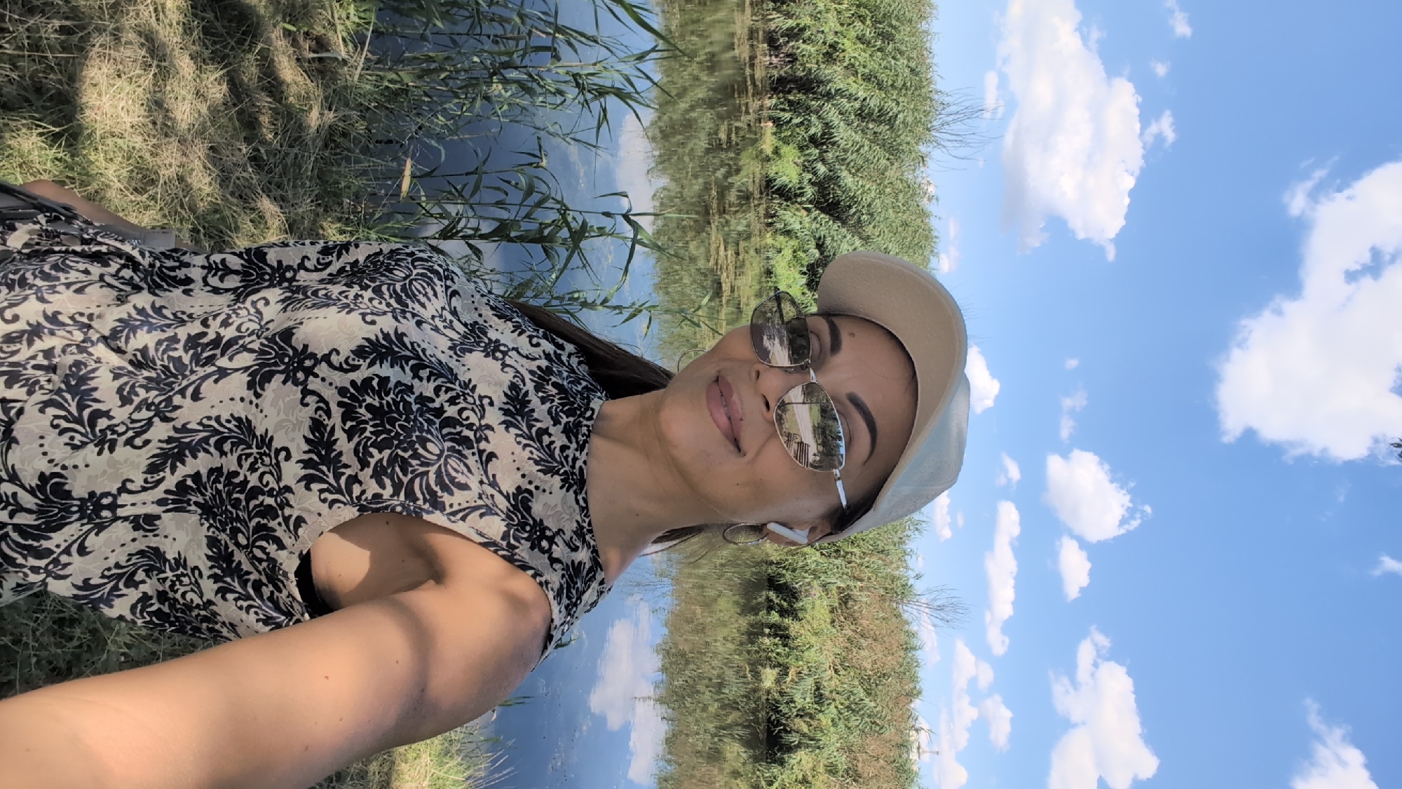 A selfie shows a woman posing outdoors with a body of water and lush green vegetation behind her. She is wearing a beige baseball cap, sunglasses, and a black and white patterned sleeveless top. She has on white earbuds and hoop earrings. The sky is bright blue with scattered white clouds. The reflection in her sunglasses shows a park bench and trees. The water appears calm, with reeds growing along the edge. The overall scene suggests a casual day out in nature, possibly in a park or nature reserve in Izmail, Ukraine, given the presence of water and dense vegetation.