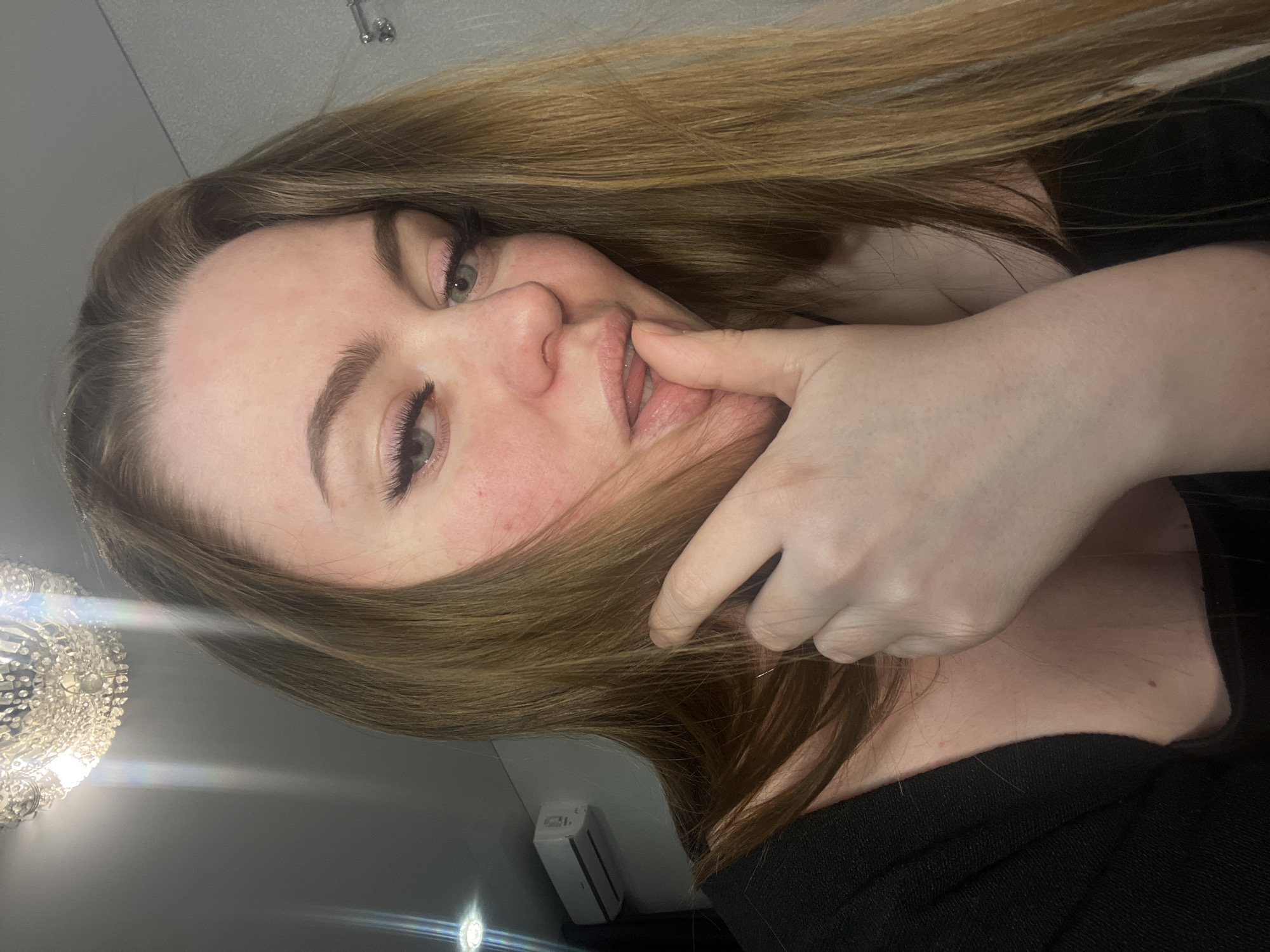 A close-up, rotated portrait shows a woman with long, light brown hair looking directly at the viewer. Her head is tilted, and her left hand is raised to her face, with fingers gently touching her lips or the side of her mouth. She has light-colored eyes, prominent eyelashes, and a soft expression, with a hint of a smile. She is wearing a dark, possibly black, top or dress.
The background consists of plain grey or off-white walls. To the left, a bright, ornate chandelier with multiple crystals is visible, acting as a strong light source and causing noticeable lens flares in the image. On the right side of the background, a white, rectangular device, possibly a wall-mounted air freshener or similar unit, is attached to the wall. The lighting is artificial and bright, indicating an indoor setting. No text is visible in the image.