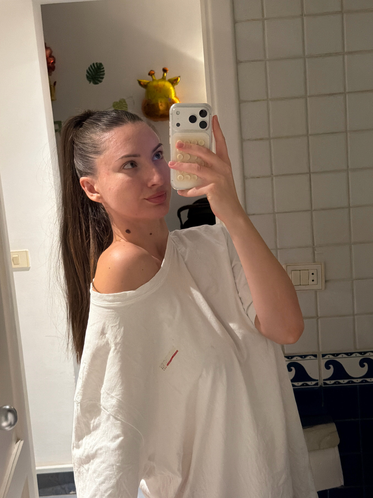 A woman with her hair in a ponytail, wearing an off-the-shoulder white t-shirt, is taking a mirror selfie in a bathroom. She is holding a white phone in her right hand. The bathroom has white tiled walls, a light switch, and a decorative band of blue and white waves above a dark blue tiled section. In the reflection on the left side of the mirror, a giraffe-shaped balloon and other decorations are visible on a wall. A doorway with a silver handle is also partially visible. The setting is consistent with a residential bathroom in Sharm El Sheikh, Egypt.