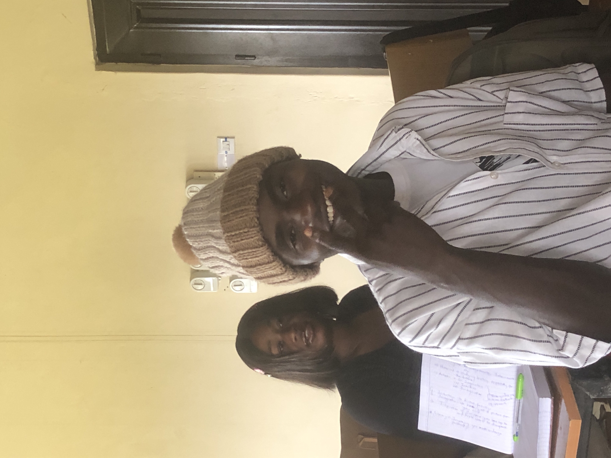 Two people are pictured, a man and a woman, in what appears to be an indoor setting. The man is in the foreground, wearing a striped shirt and a knitted beanie. He is smiling and looking towards the camera. The woman is behind him, wearing a black top, and also smiling. They are both looking at the camera. A notebook with some writing and a green pen are visible on a desk in front of them. The wall behind them is a pale yellow color, and there are light switches on the wall. The image has been rotated, with the subjects appearing sideways. The lighting suggests it is daytime. There is no visible text in the image. The overall mood seems cheerful and relaxed.