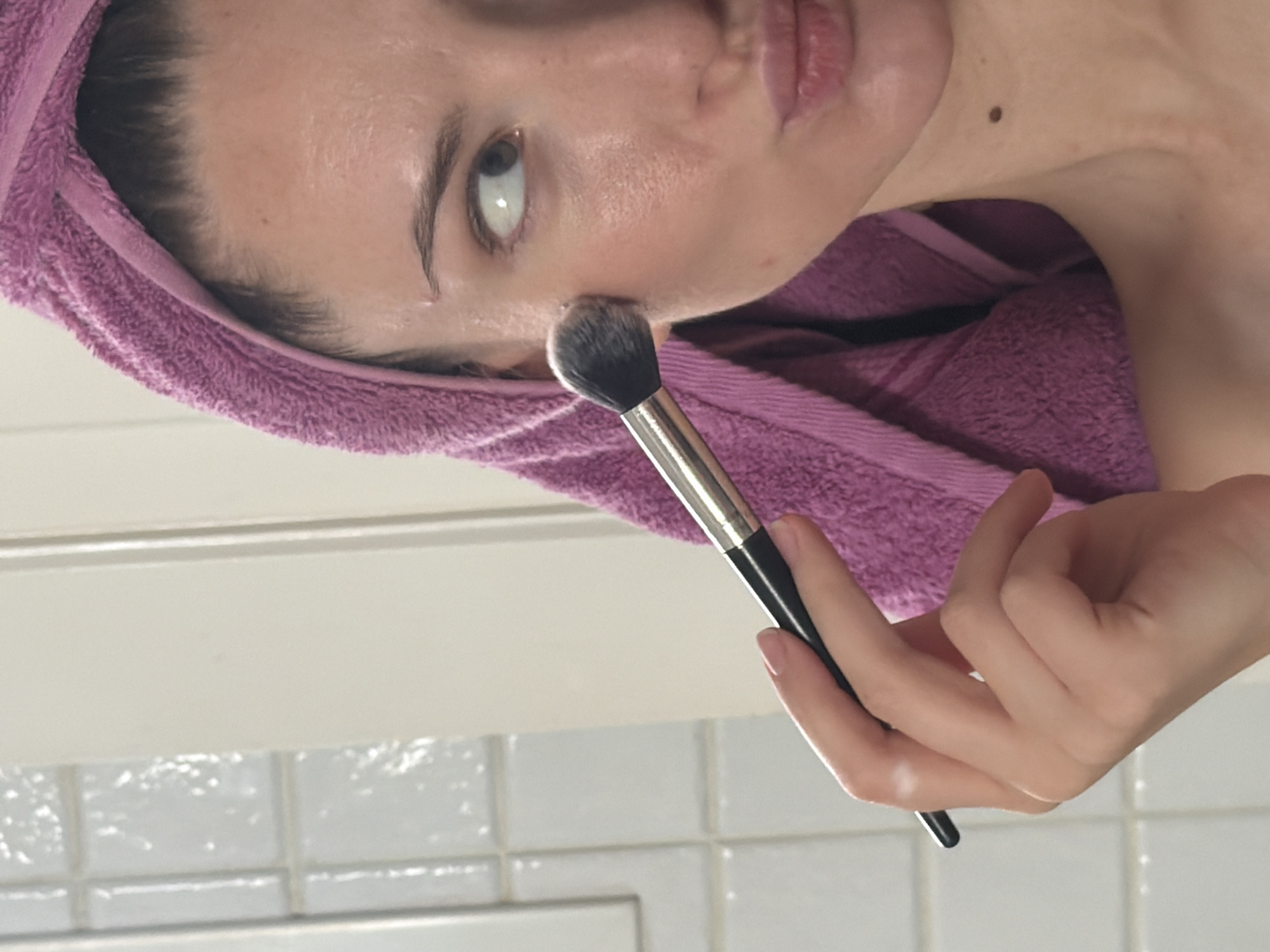 A woman with a purple towel wrapped around her head is applying makeup to her cheek with a brush. Her head is angled to the right, and her eyes are looking to the right. The background appears to be a tiled wall. The woman's skin appears slightly wet, suggesting she may have recently washed her face. There is a mole on her neck. The image does not contain any information that would allow for the determination of its location in Sharm El Sheikh, Egypt.