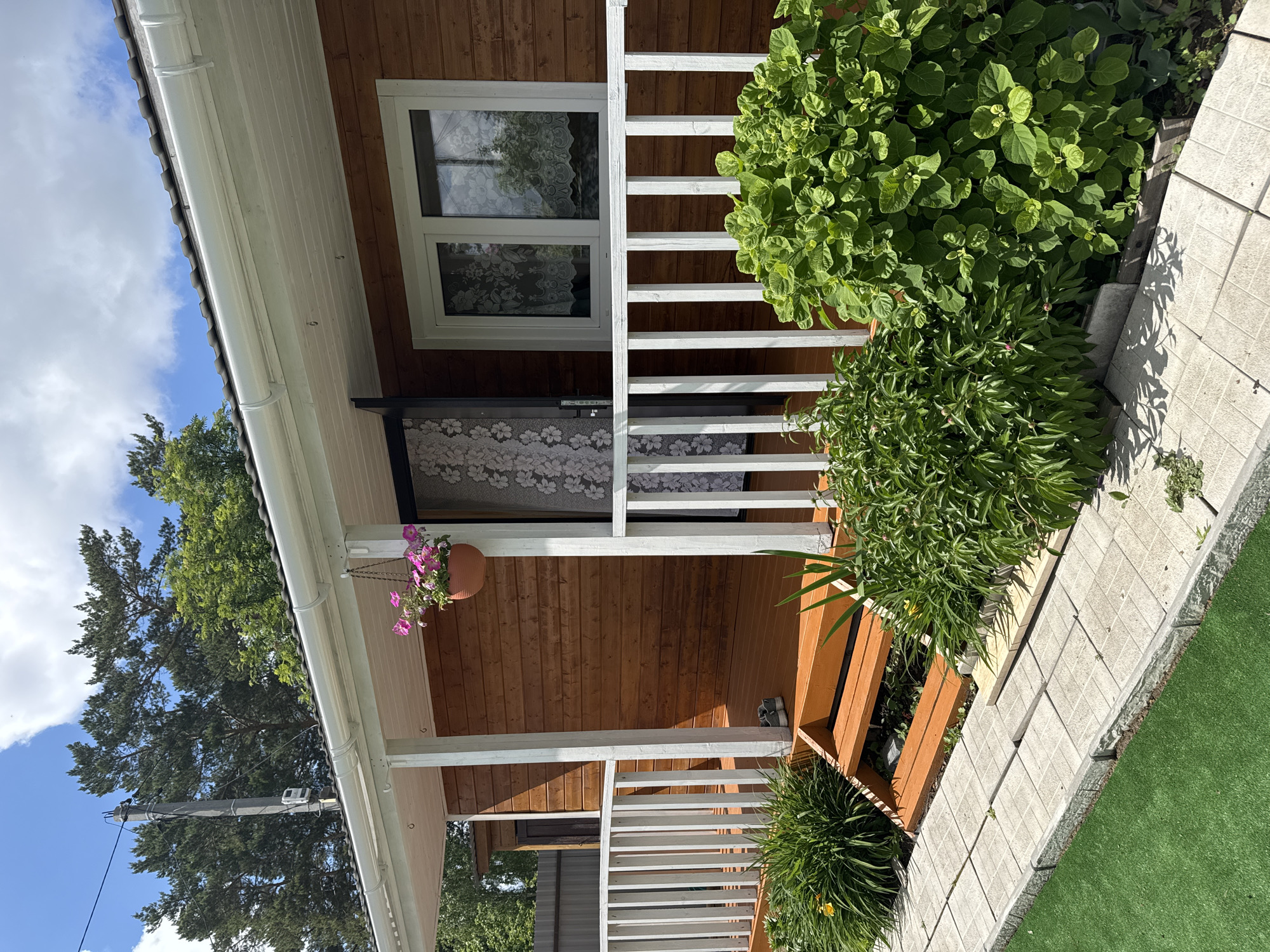 The image shows a section of a wooden building, possibly a house or cabin, with a small porch area. The building is constructed of horizontal wooden planks, stained a medium brown color. A window with white trim is visible, covered by a floral-patterned curtain. The porch has a white railing. A hanging flower pot with pink flowers is suspended from the roof.
To the right of the porch, there is a landscaped area with green foliage and a stone walkway. A patch of artificial green turf is also visible.
A power pole can be seen in the background on the left.
No people are visible.
Based on the location information provided, this scene would be found in Chaikovsky, Russia.