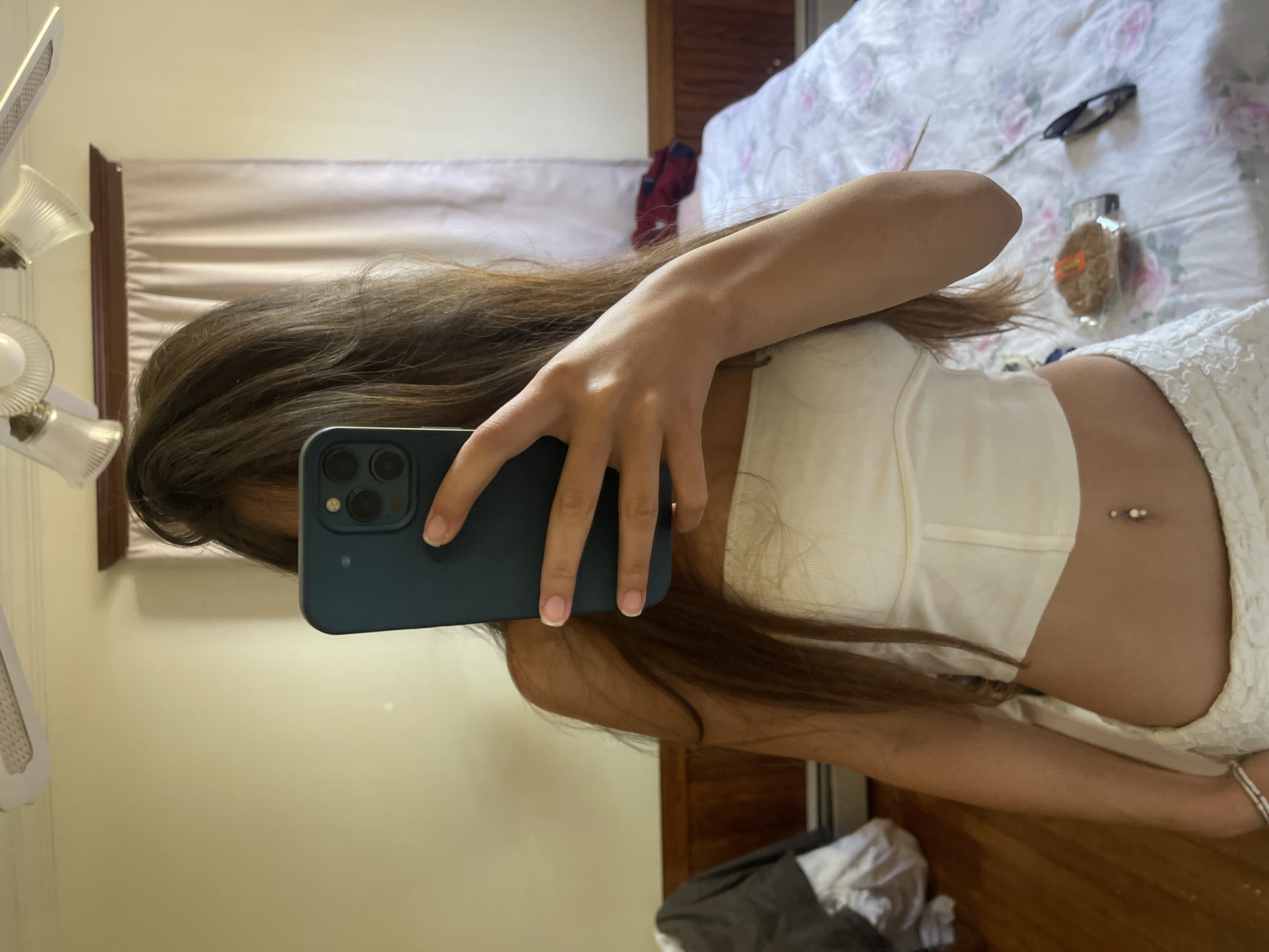 A young woman is standing in a bedroom, taking a mirror selfie. She is wearing a white strapless top and white skirt, and has a belly button piercing. She is holding a dark blue smartphone in her right hand, covering her face. The room has light yellow walls, a ceiling fan, and a wooden floor. A bed with a floral patterned comforter is visible in the background, along with a closet and a window with light-colored curtains. There is also a piece of packaged food on the bed. The location is Oak Bluffs, United States of America.
