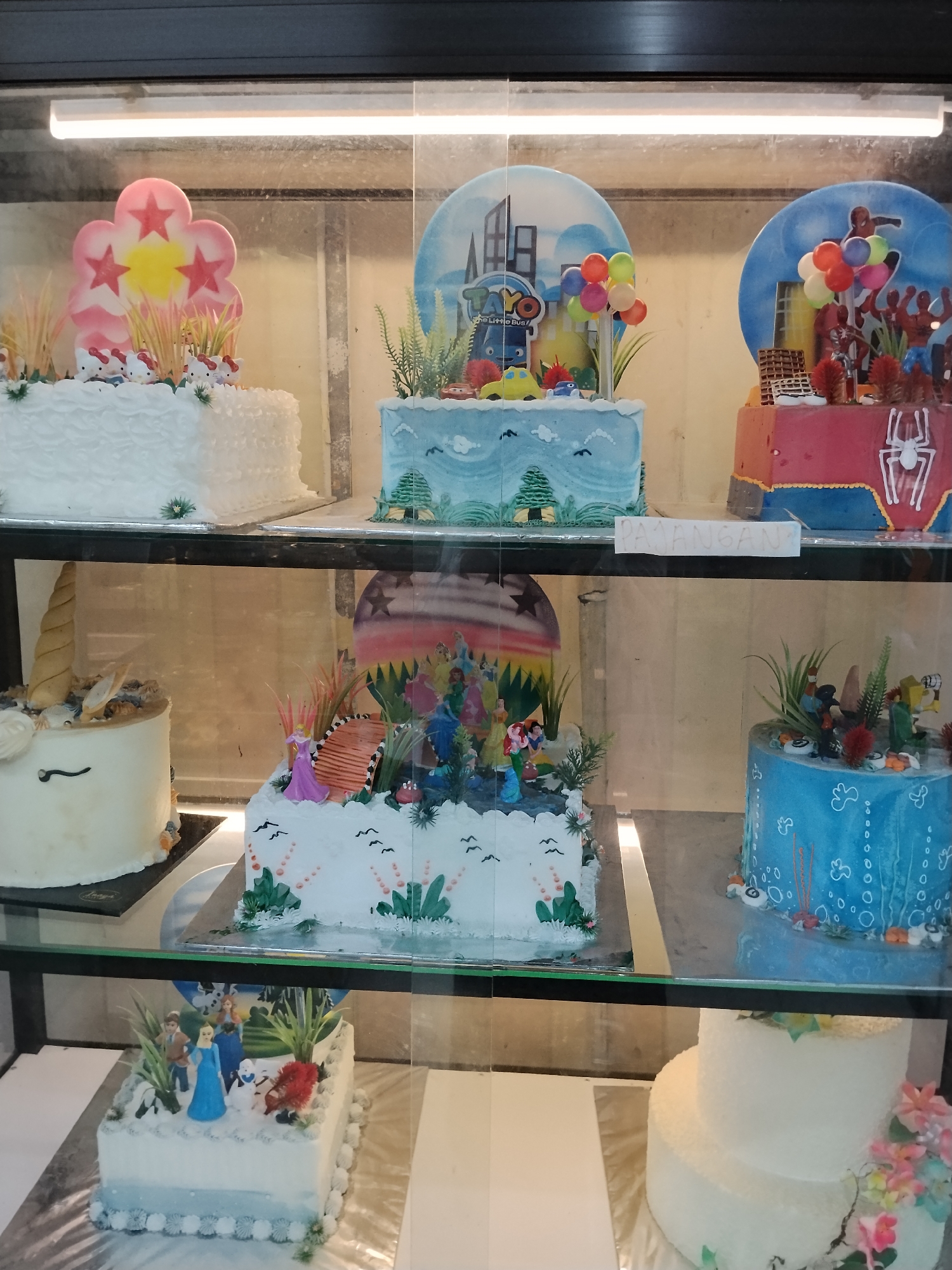 In a bakery display case in Ciruas, Indonesia, a variety of decorated cakes are presented. The top shelf features three cakes: a white cake topped with pink and yellow flower decorations and Hello Kitty figures, a blue cake with a backdrop depicting cartoon characters and balloons, and a red cake with a Spiderman theme. The middle shelf holds a unicorn-themed cake on the left, a white cake decorated with princess figures and greenery in the center, and a blue cake adorned with aquatic imagery on the right. The bottom shelf displays a rectangular white cake featuring animated character figurines, and a multi-tiered white cake decorated with flowers on the far right. The cakes are illuminated by overhead lighting.