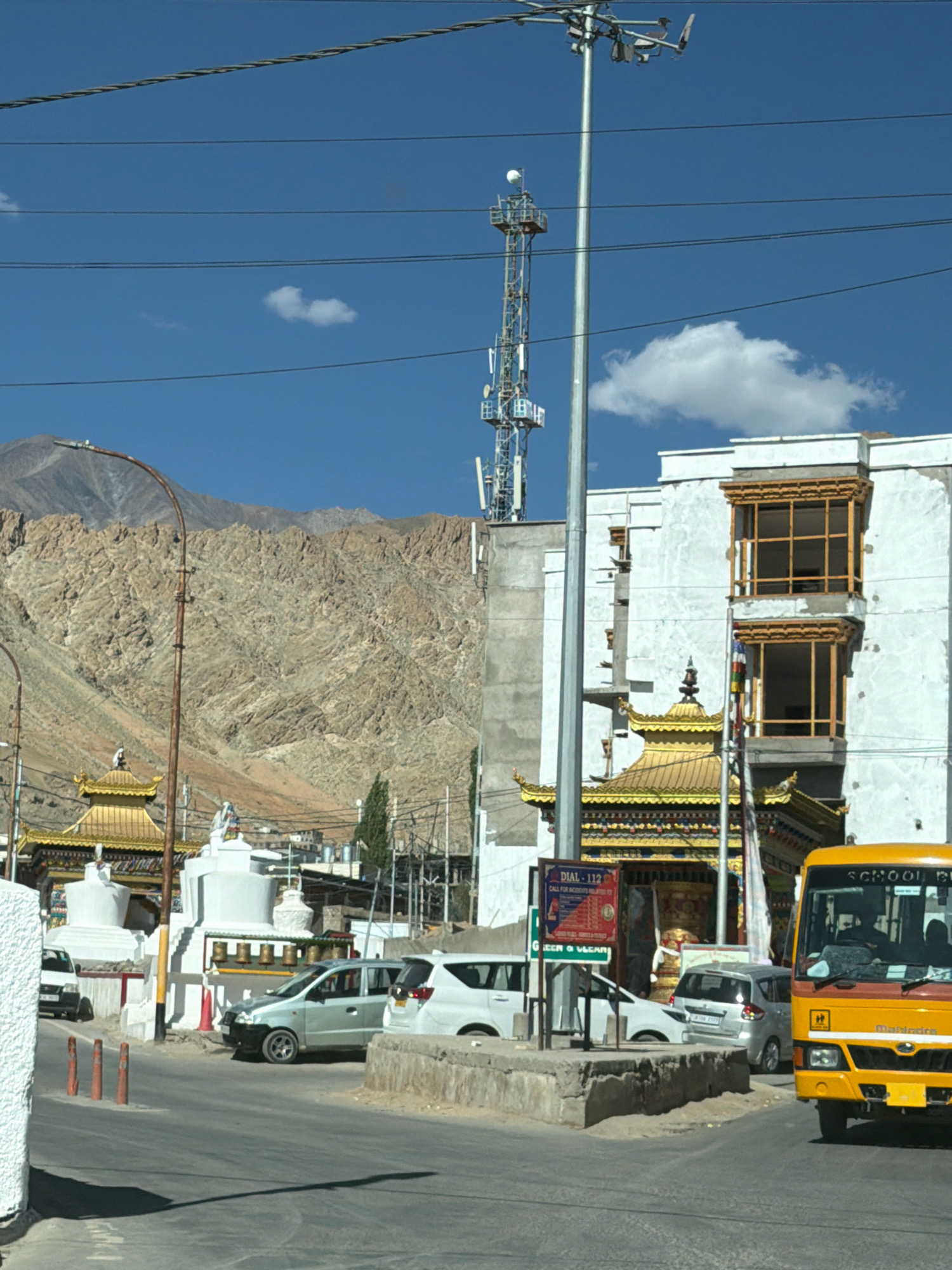 In the city of Leh, India, a bright yellow school bus dominates the foreground, traveling on a paved road. To the left of the bus, a light silver car is parked, and another silver car is driving on the road. In the background, several white stupas and a building with a golden roof are visible, hinting at a religious or cultural site. Behind these structures, a large, barren mountain looms under a clear blue sky, dotted with a few wispy clouds. A tall cell tower stands prominently in the middle ground, alongside power lines stretching across the frame. The scene captures a typical day in Leh, with a blend of modern infrastructure and traditional architecture set against a dramatic natural landscape.