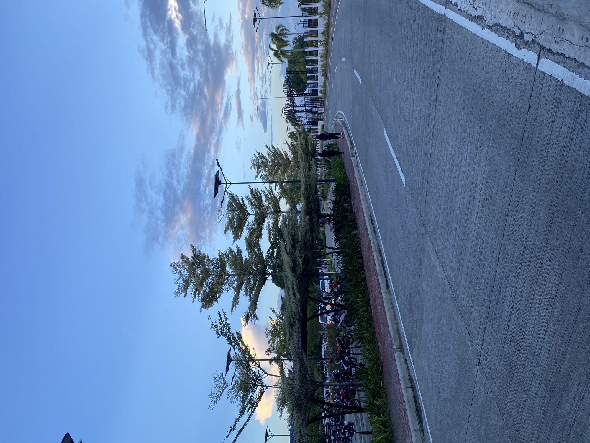 The image depicts a paved road curving to the right, with a sidewalk and a row of trees on the left. The sky is a gradient of blue with scattered clouds, suggesting either early morning or late afternoon. Streetlights with solar panels are visible along the road. A parking area with motorcycles and some cars is situated behind the trees. In the background, a decorative gate and palm trees are visible, characteristic of tropical environments. The scene appears to be a well-maintained outdoor area, possibly a park, entrance to a complex, or a promenade. Based on the architecture and vegetation, this location is likely in Davao City, Philippines. There are two individuals walking on the sidewalk in the middle ground of the image, facing away from the camera. The overall impression is one of a serene, urban landscape at dusk or dawn.