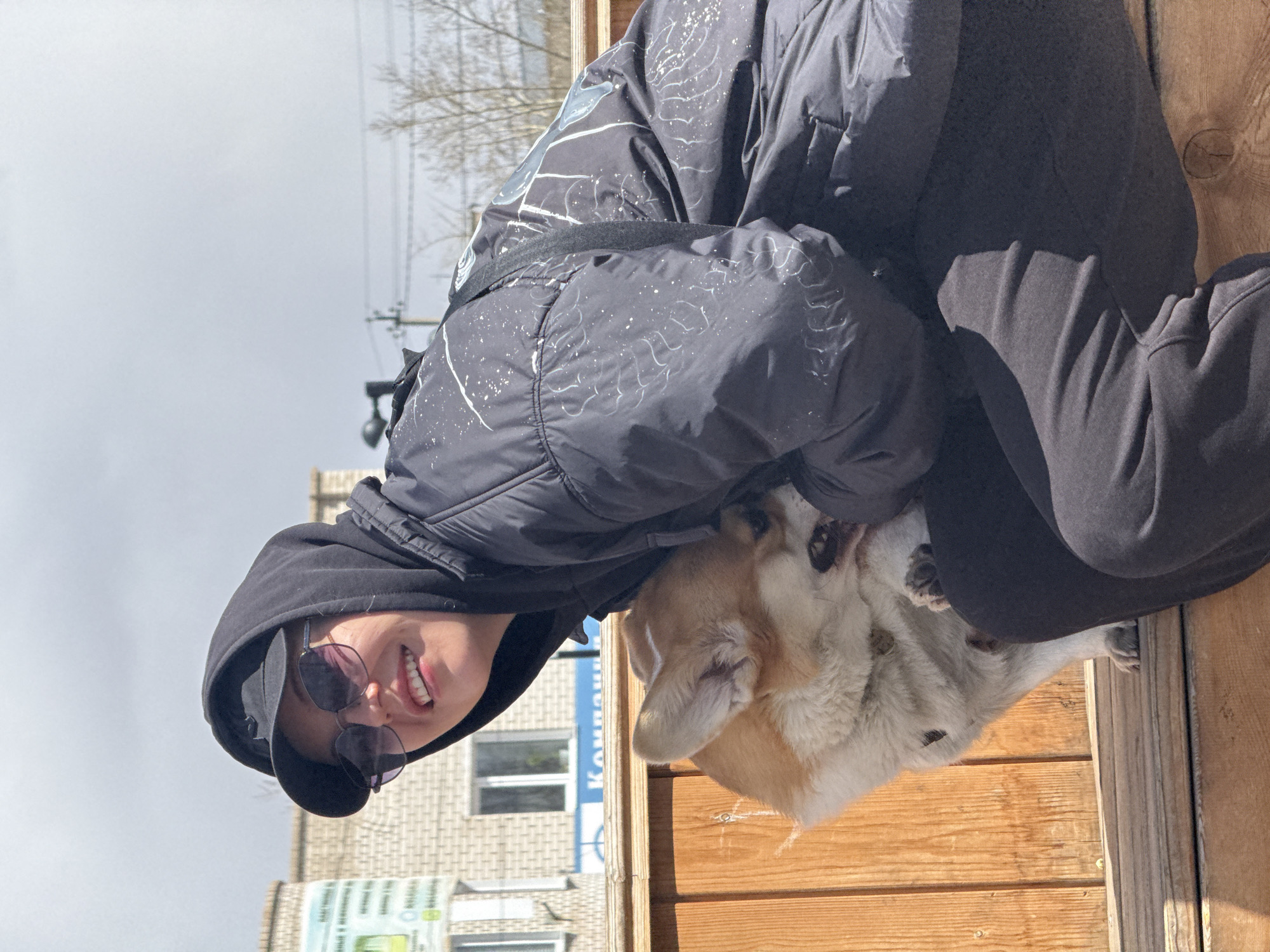 A person in a black hooded jacket and sunglasses is kneeling and hugging a tan and white dog. The person is smiling. The background includes a light-colored brick building and bare trees, suggesting an urban environment. A wooden structure, likely a deck or wall, is in the foreground. The signage in the background contains Cyrillic script, indicating a location in Russia, confirmed to be the city of Borzya.