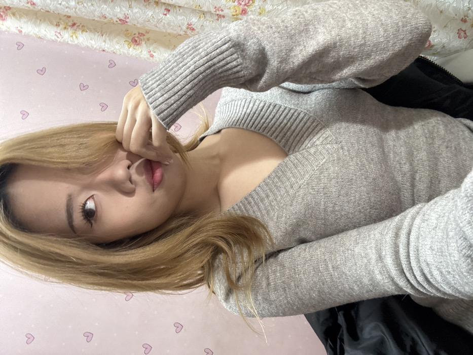 A woman with blonde hair wearing a gray V-neck sweater poses for a close-up shot. Her left hand touches her face near her lips. The background consists of a pink wall adorned with small heart patterns and a floral-patterned headboard or curtain. The lighting is soft, highlighting her skin and hair color. A black garment is visible behind her right shoulder. The image is rotated counter-clockwise.