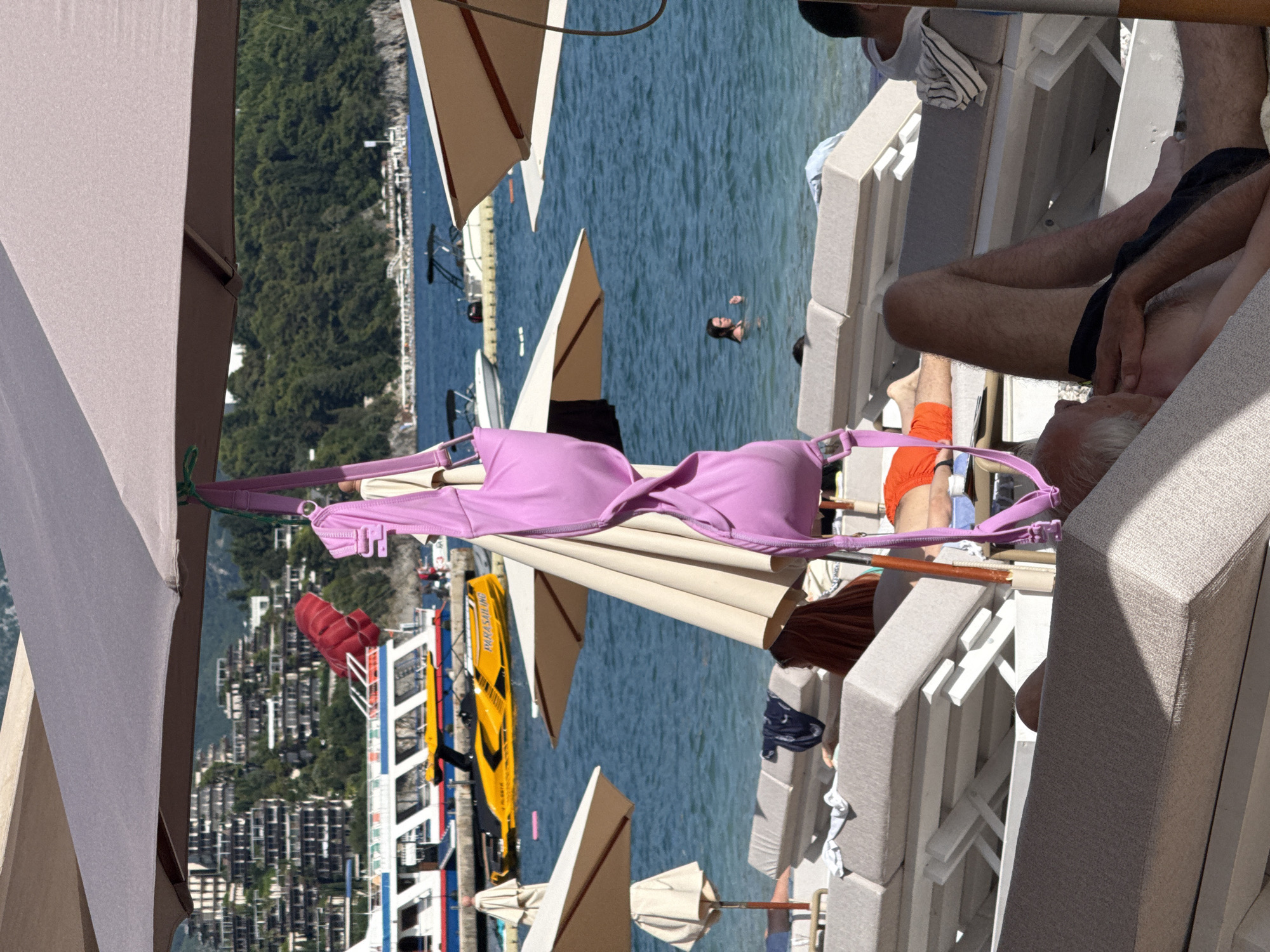 The image captures a sunlit beach scene in Budva, Montenegro. In the foreground, numerous tan-colored sun loungers are arranged on a pebble beach, partially shaded by large, open beach umbrellas. An older man with gray hair, bare-chested and wearing dark swim trunks, lies on a central lounger, facing upward. To his right, another person in orange swim shorts is visible on a lounger, facing away. Further back on the left, a person with reddish-brown hair is also reclined on a lounger.
A prominent pink bikini top hangs from the pole of a gray beach umbrella in the center of the frame. Below it, a beige umbrella is also open.
The mid-ground features calm, blue sea water with several boats. A large white and blue passenger boat is docked on the left, next to a yellow "PARASAILING" boat, which has a deflated red parachute visible behind it. Further out, smaller speedboats and leisure crafts are moored or moving. A person with dark hair is visible swimming in the water on the mid-right.
In the background, lush green, tree-covered hills rise, dotted with distinctive terraced white buildings that ascend the slopes. Another distant coastline with faint structures is visible across the bay on the far right. The sky is partially obscured by the umbrellas in the foreground.