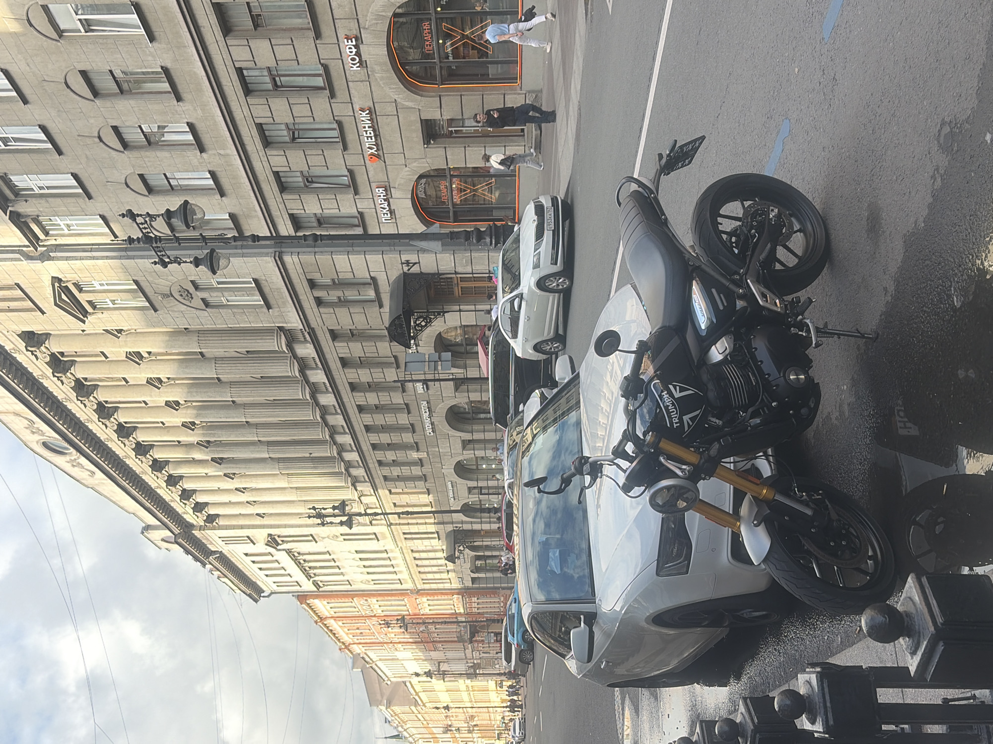 The image depicts a street scene in Saint Petersburg, Russia, under an overcast sky, suggesting a cool or perhaps rainy day. The primary subjects are a parked motorcycle and a white car, both situated on a wet asphalt road. The motorcycle, a Triumph, is positioned in the foreground, angled towards the camera, with its front wheel turned. It is dark in color with gold accents on the front forks. The white car is parked behind it, taking up a significant portion of the right side of the frame.
The background is dominated by grand, ornate buildings characteristic of Saint Petersburg architecture. These buildings feature multiple stories with numerous windows and classical decorative elements, including columns and intricate cornices. Along the street, several lampposts are visible, adding to the urban ambiance.
There are a few people visible in the distance, walking along the sidewalk or crossing the street. Their presence suggests a normal day of activity in the city. Signs with Cyrillic text are visible on the buildings, indicating shops or businesses. One sign reads "КОФЕ" (coffee), another "ХЛЕБНИК" (baker), and "ПЕКАРНЯ" (bakery). The wet road surface and the cloudy sky imply recent rain or ongoing damp weather. The scene feels like a typical urban environment captured during daylight hours.